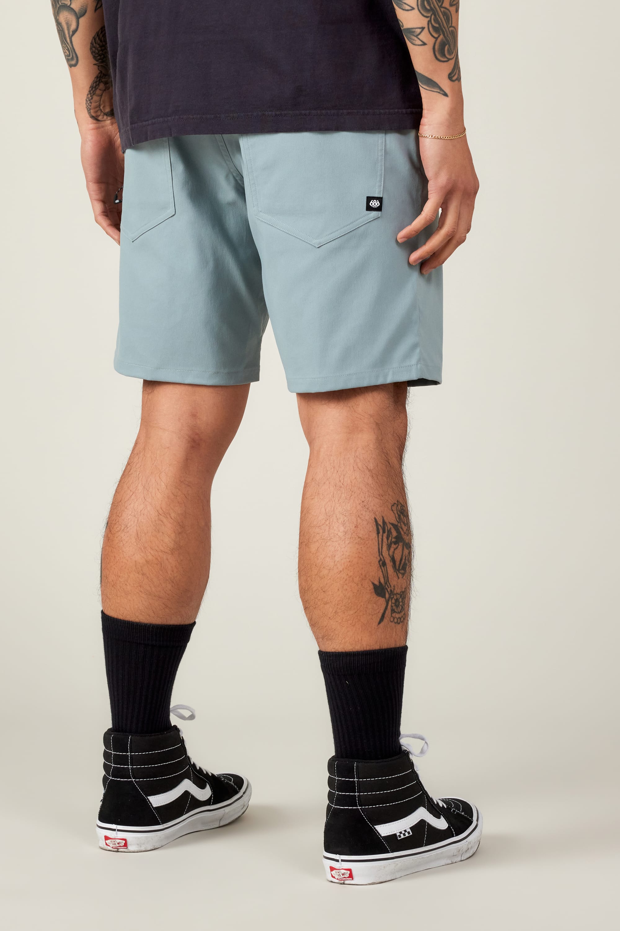 A person stands facing away, wearing light blue 686 Everywhere® Hybrid Shorts—breathable and stain-resistant—paired with black socks, black Vans sneakers, and a dark T-shirt. Their tattooed arms and legs stand out against the off-white background.