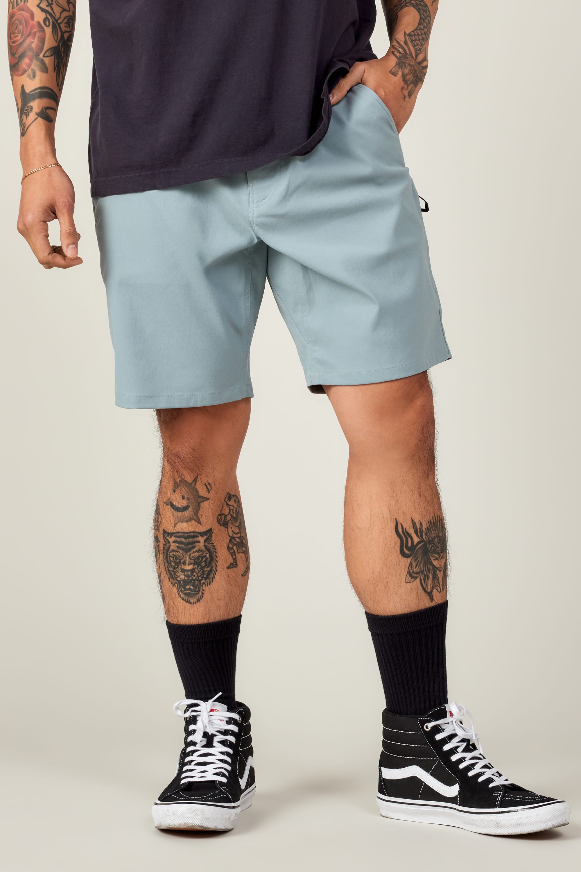A person wearing light blue 686 Everywhere® Hybrid Shorts, a dark t-shirt, black socks, and black high-top sneakers. Tattoos are visible on both arms and legs, with their left hand in their pocket. The background is plain.