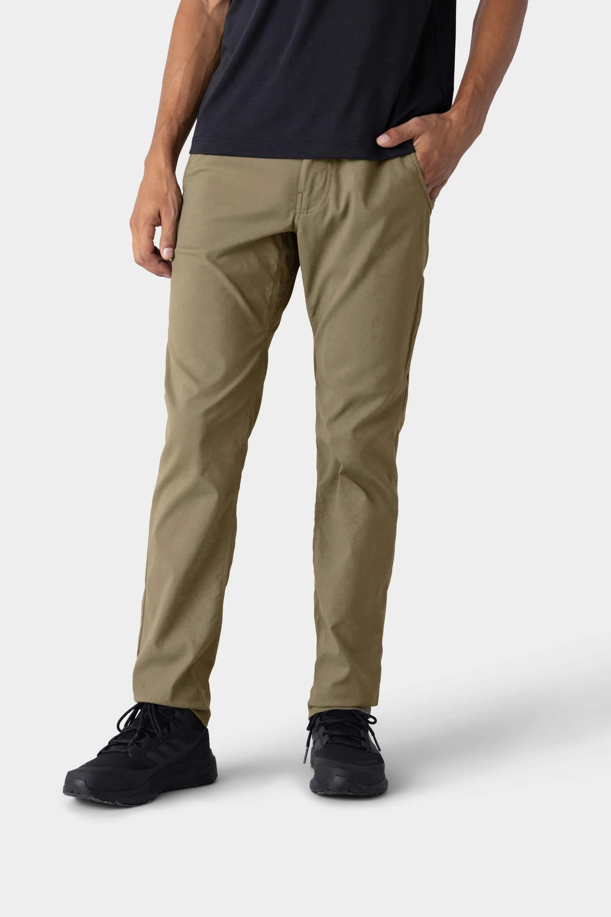 A person wears 686 Men's Everywhere® Merino-Lined Pant - Slim Fit in khaki, paired with a black t-shirt and black shoes, standing against a plain light gray background with one hand in their pocket, dressed for colder weather.