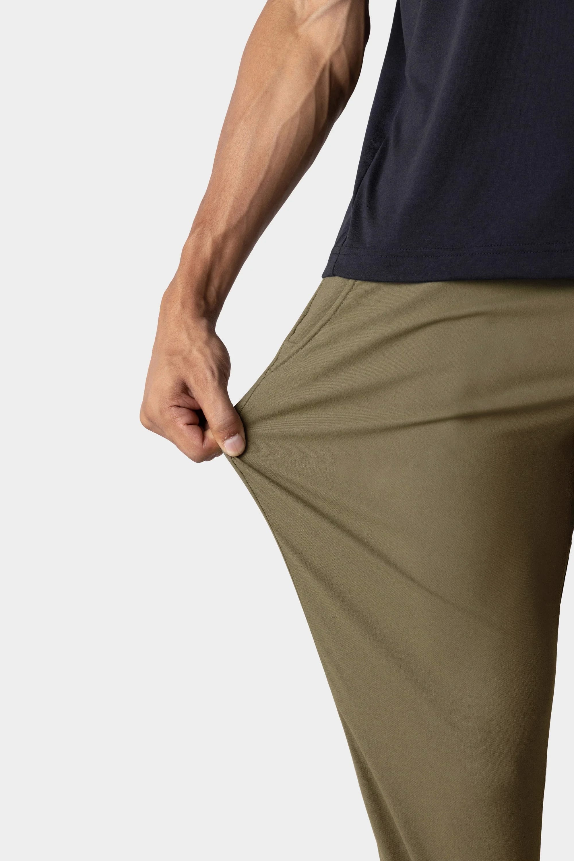 A person in a black shirt stretches the fabric of their olive green 686 Men's Everywhere® Merino-Lined Pant - Slim Fit, showing its flexibility and warmth against a plain white background.