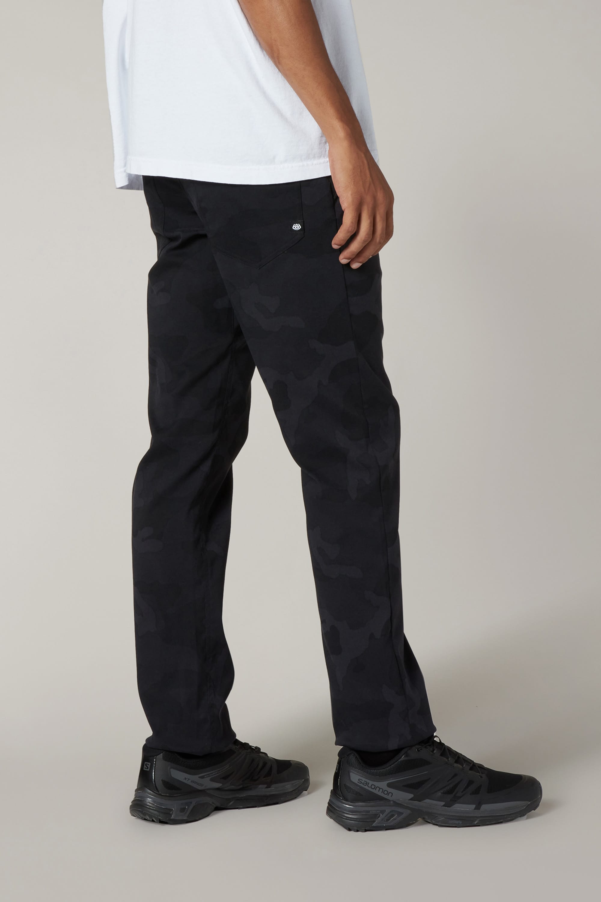 A person stands against a plain background, showing only the lower half, wearing 686 Men's Everywhere® Pant - Slim Fit in black camo and black sneakers with a white shirt.