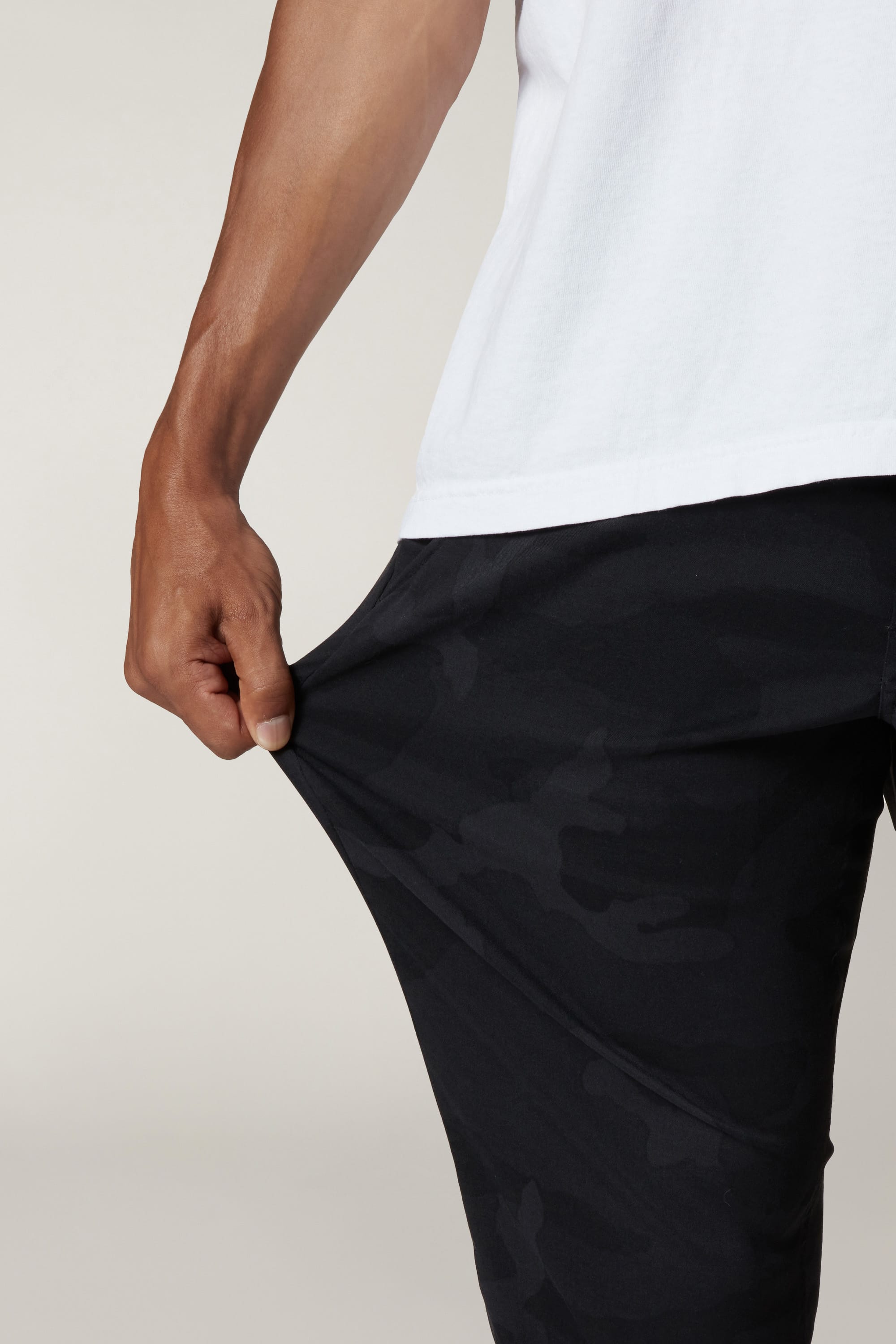 A person in a white shirt stretches the fabric of their dark, camo-patterned 686 Men's Everywhere® Pant - Slim Fit, highlighting flexibility. Only the lower torso and one arm are visible against a plain background.