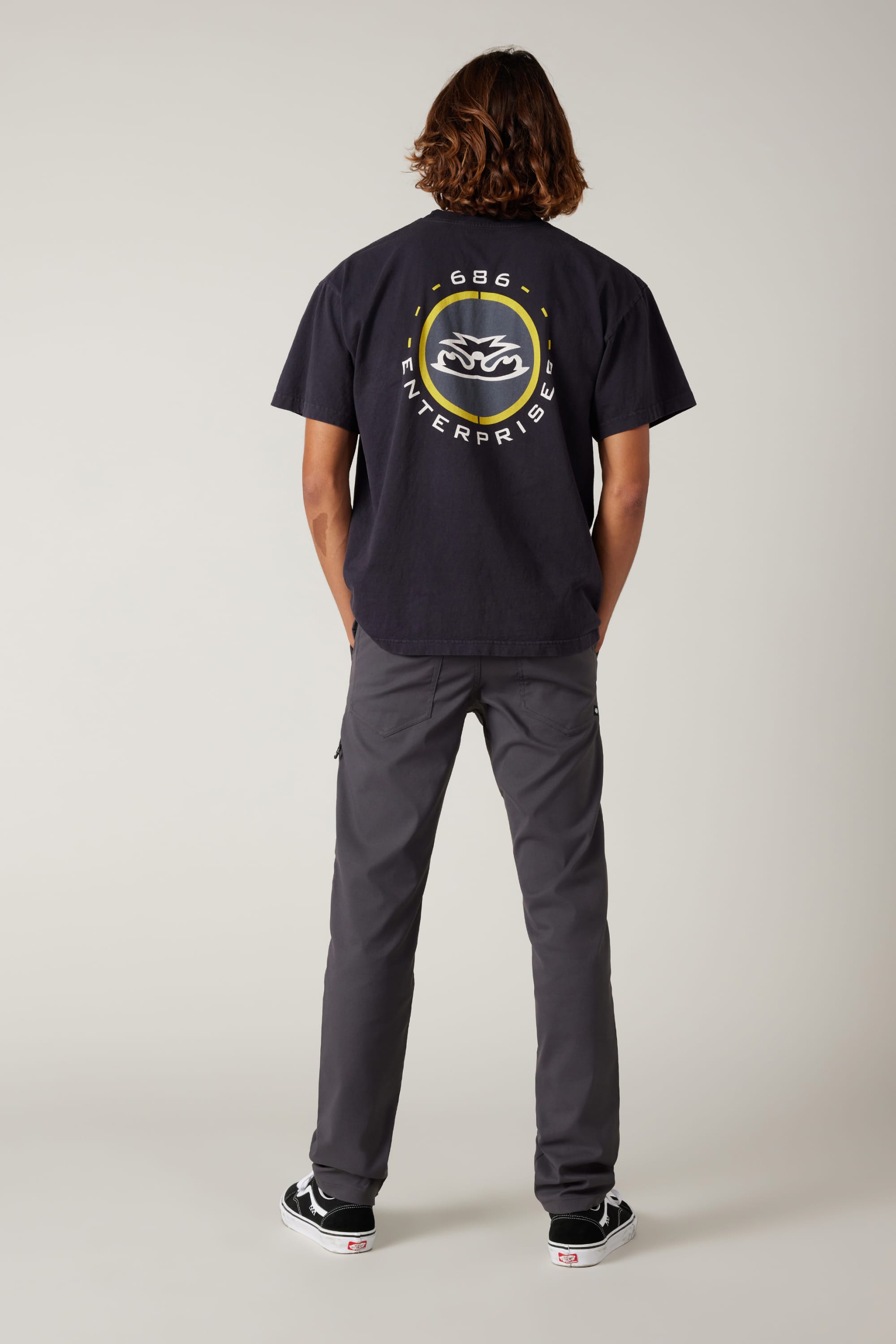 A person with shoulder-length hair faces away, wearing a black 686 graphic tee, 686 Men's Everywhere® Pant - Slim Fit in gray, and black-and-white sneakers, set against a plain light gray background.