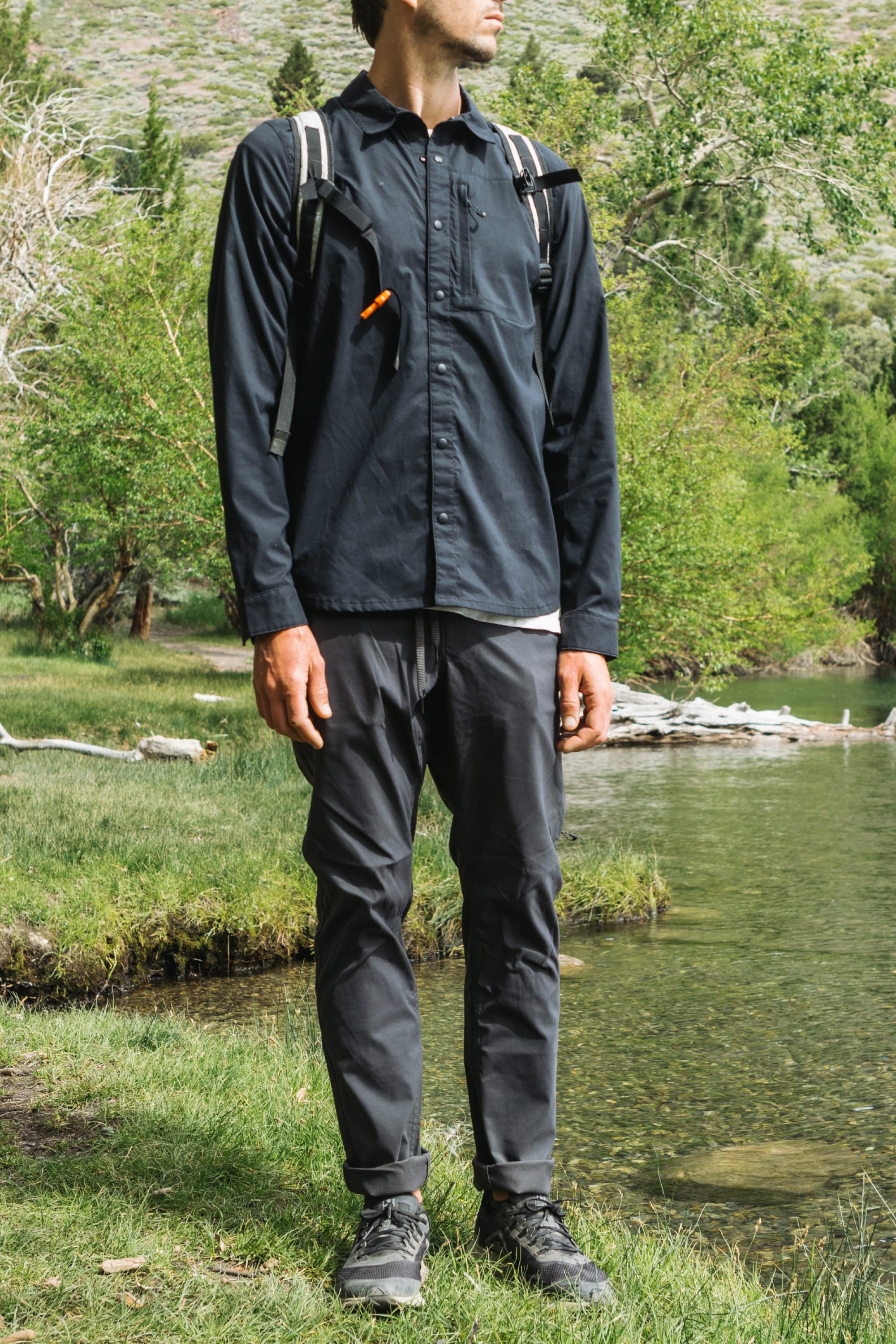 A person in a dark long-sleeve shirt, 686 Men's Everywhere® Pant - Slim Fit, hiking boots, and a backpack stands on grass by a lake with trees and hills behind them. Their face is not fully visible.