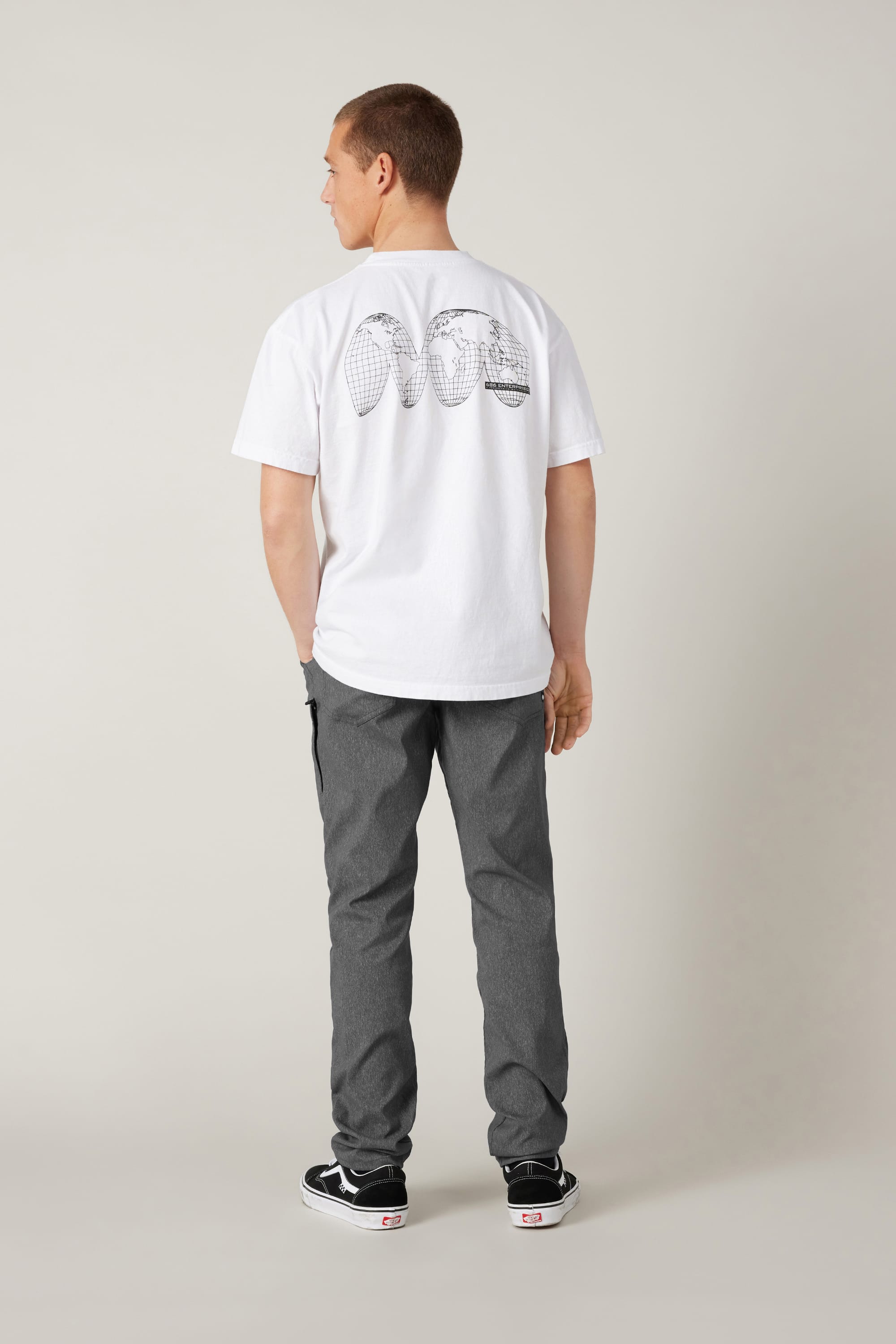 A man stands facing away, wearing a white T-shirt with a black graphic on the back, grey 686 Men's Everywhere® Pant - Slim Fit, and black Vans sneakers, against a plain light-colored background.