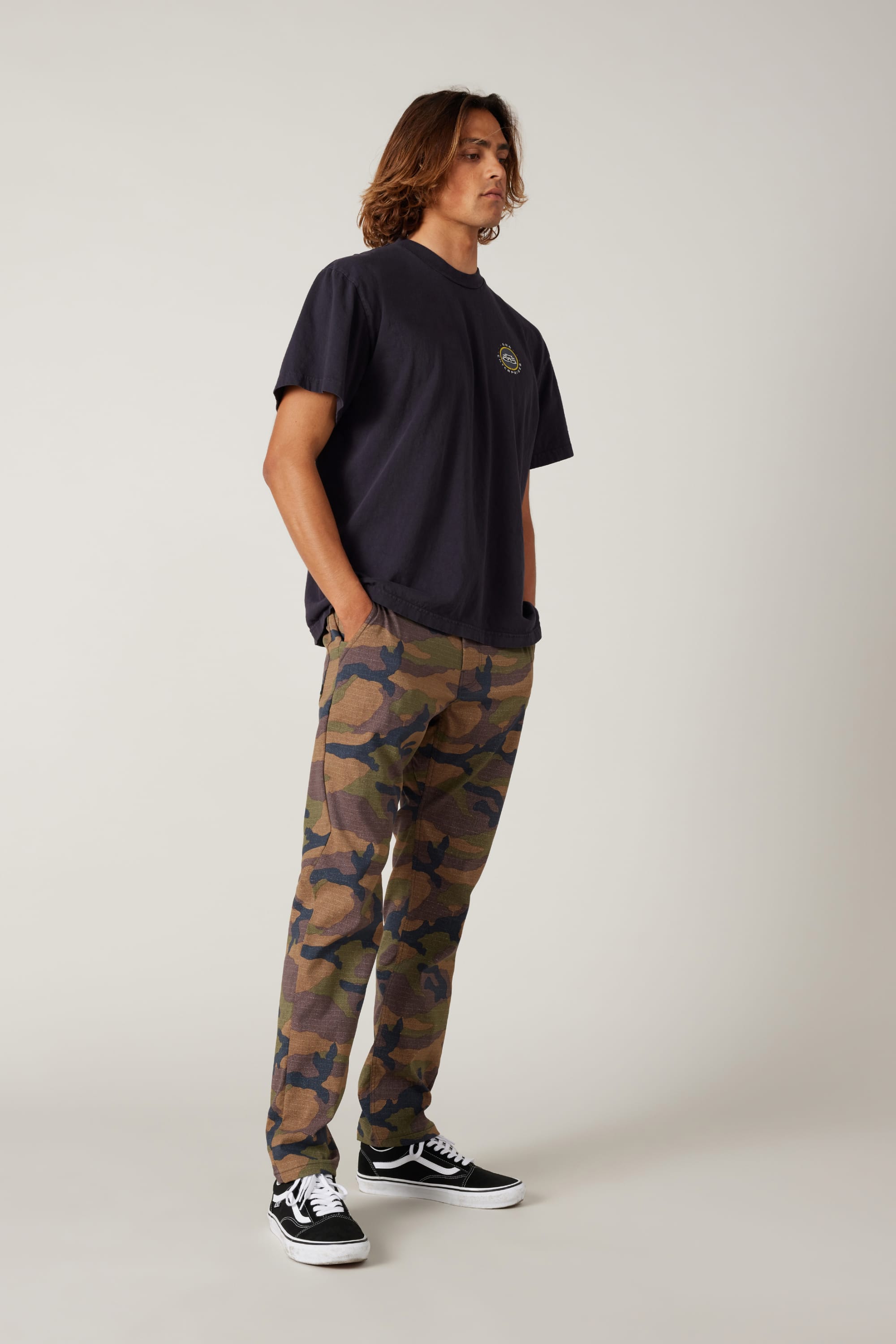 A person with long hair wears a loose black T-shirt, 686 Men's Everywhere® Pant - Slim Fit, and black Vans sneakers. They stand against a plain, light background with hands in their pockets, looking slightly to the side.