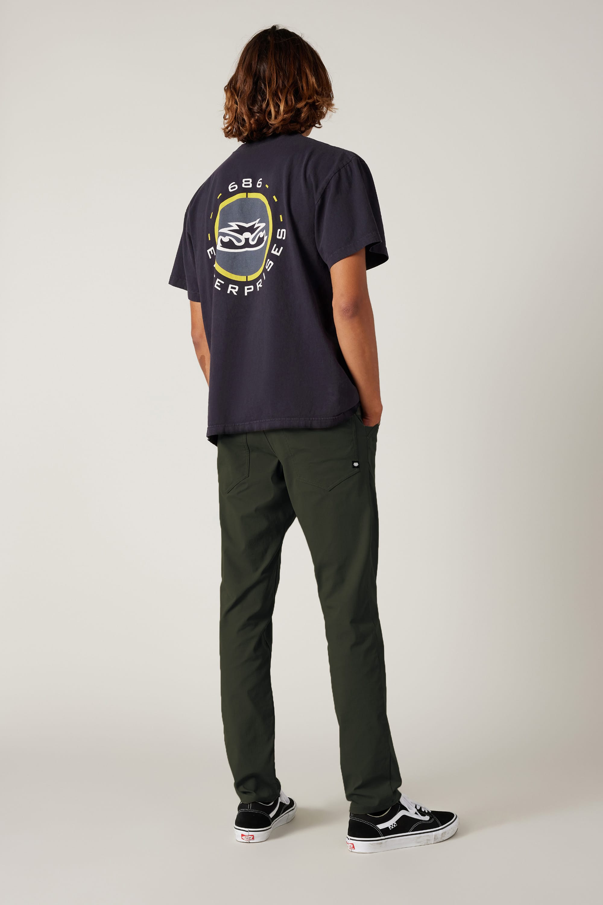 A person with shoulder-length hair faces away, wearing a dark graphic tee, 686 Men's Everywhere® Pant - Slim Fit in olive green, and black Vans sneakers, against a plain light background.