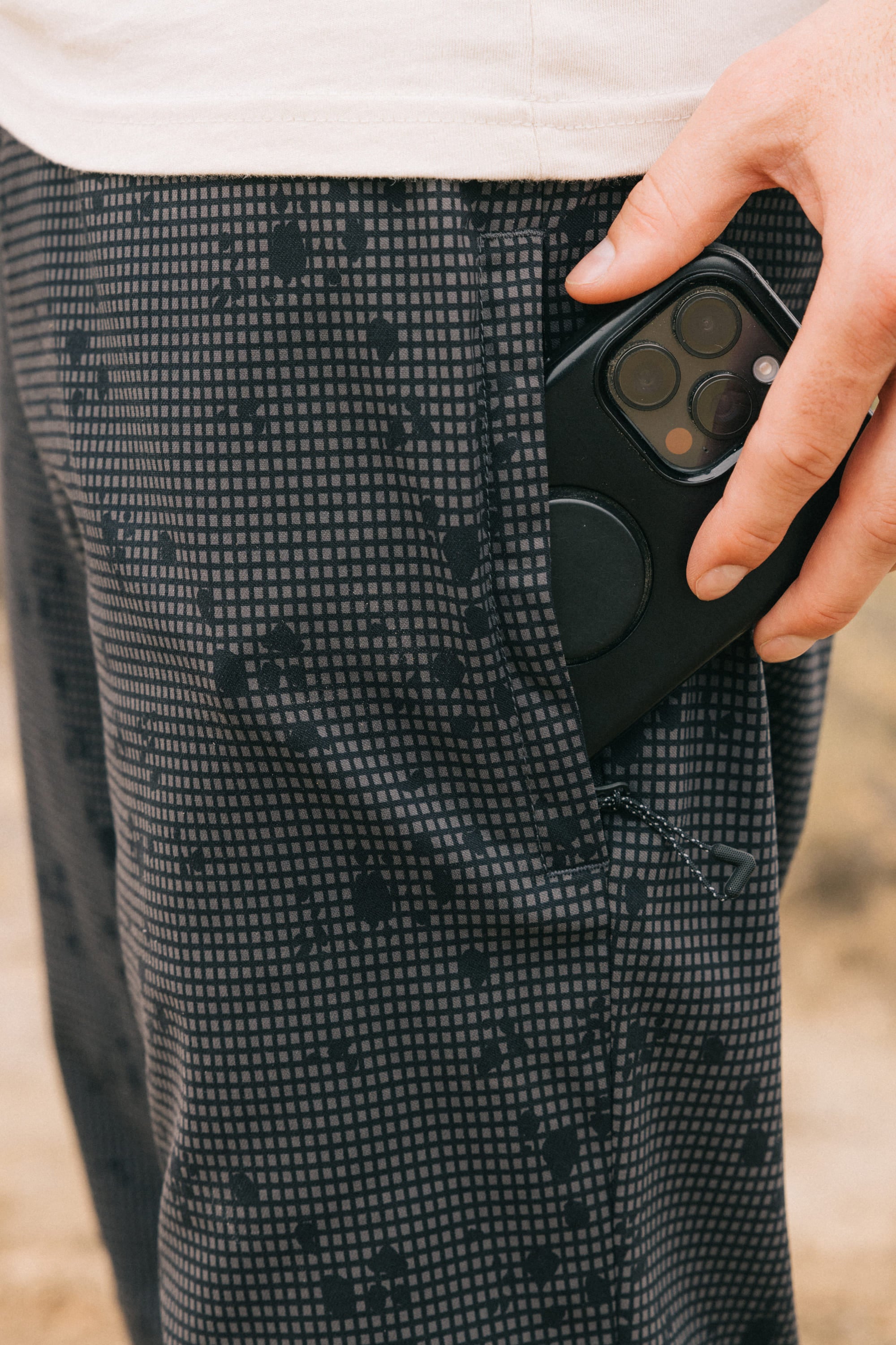 A hand places a black smartphone with a circular grip into the pocket of gray, breathable 686 Men's Everywhere® Pant - Slim Fit. The wearer has a white shirt, and the background is blurred.
