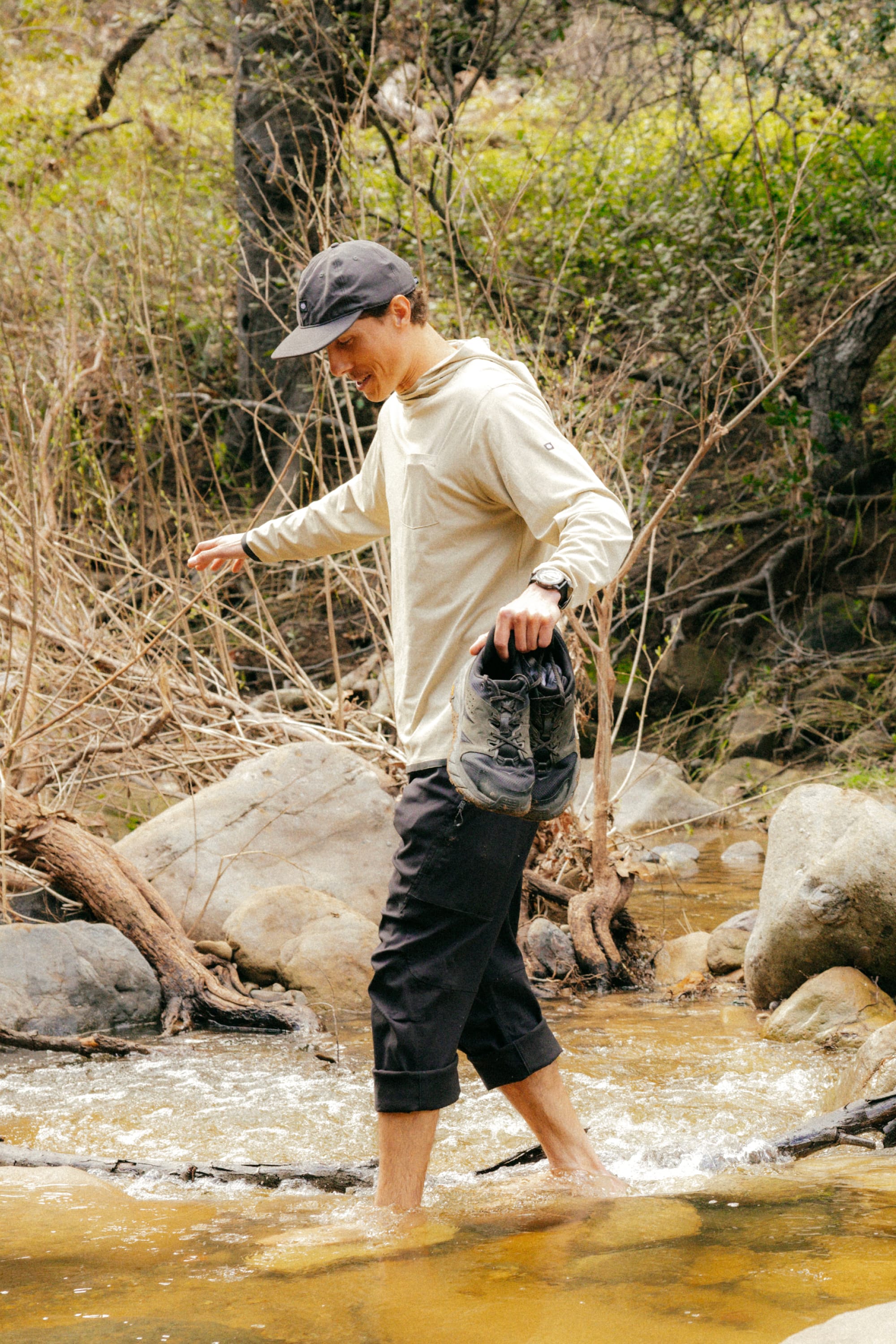 A person wearing a cap, long-sleeve shirt, and 686 Men's Anything Cargo Pant - Relaxed Fit by 686 walks barefoot through a rocky creek with shoes in hand, surrounded by trees and vegetation.