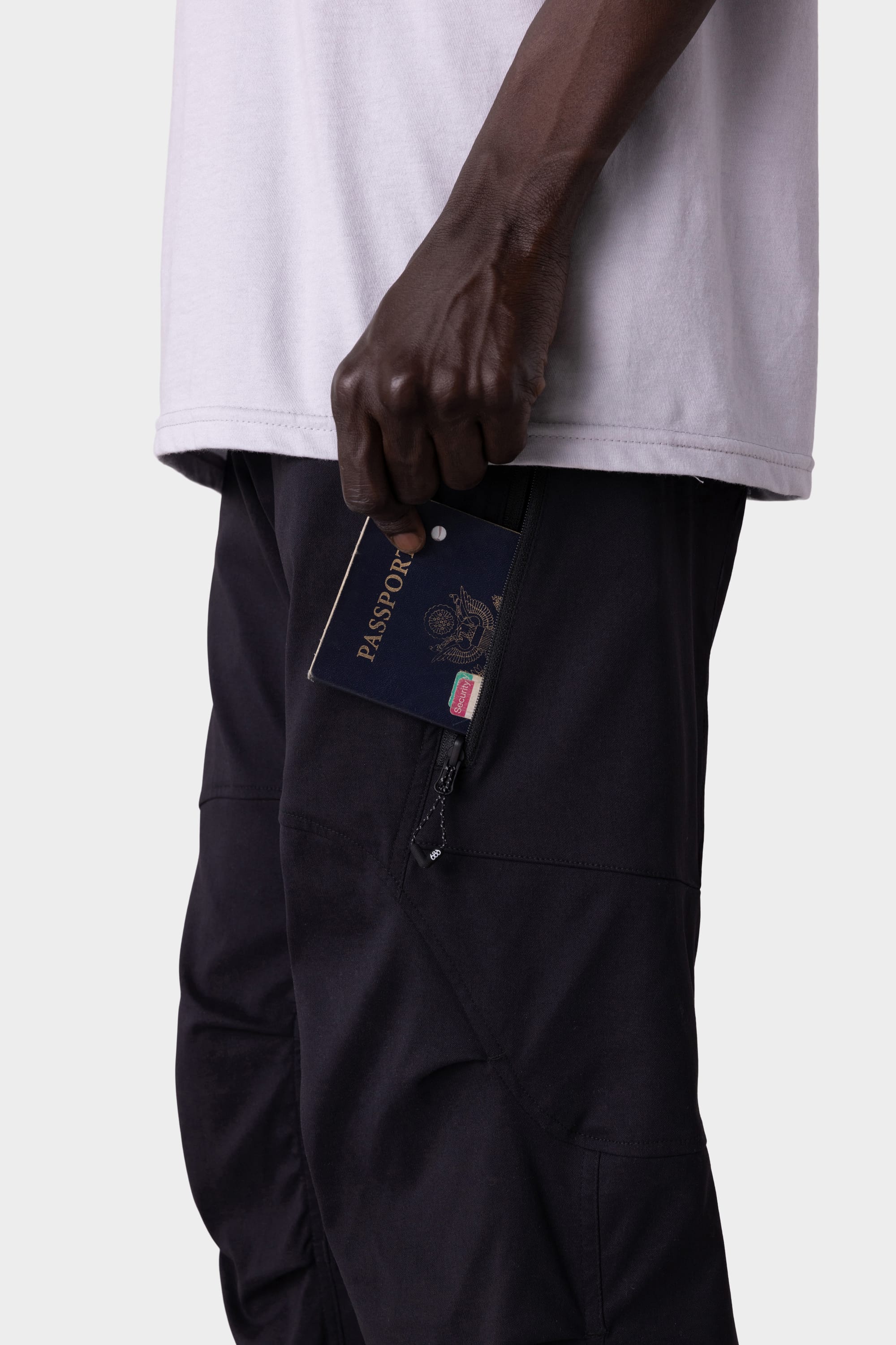 A person wearing a white shirt and 686 Men's Anything Cargo Pant - Relaxed Fit holds a passport partly tucked into a zippered side pocket. The close-up photo highlights the hand and pocket area against a plain background.