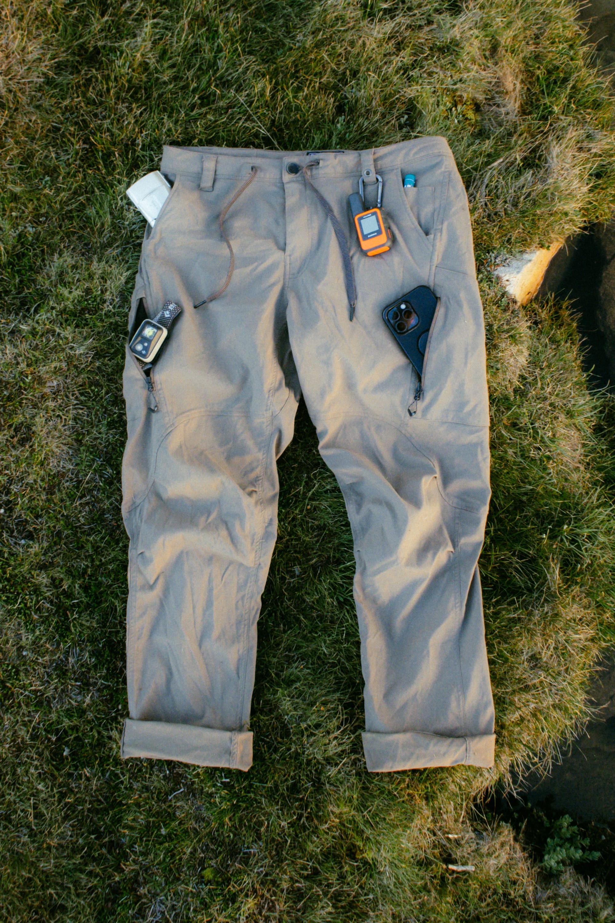 A pair of 686 Men's Anything Cargo Pant - Relaxed Fit in light brown lies on grass, with a GPS device, smartphone, and compass tucked into its 13 pockets and waistband.