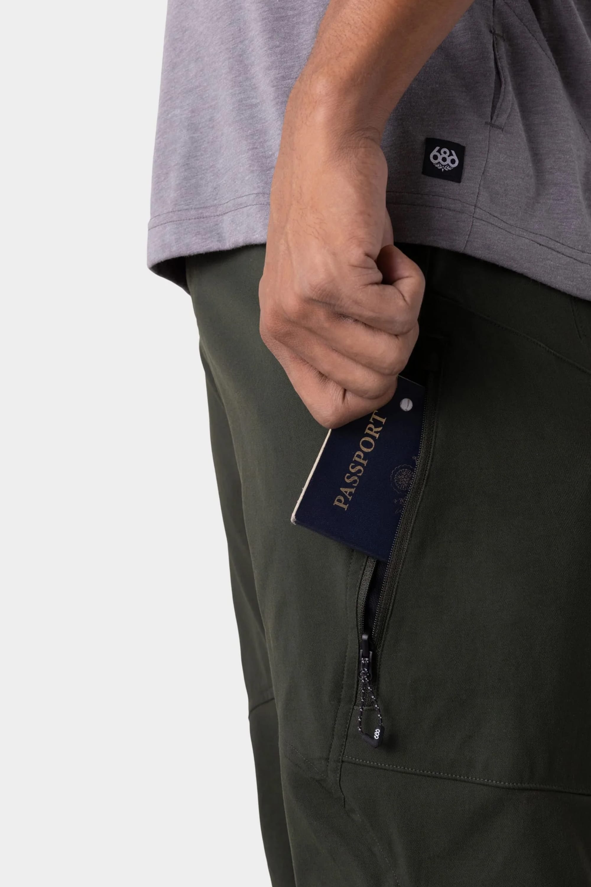 A person in a gray shirt and 686 Men's Anything Cargo Pant - Relaxed Fit by 686, featuring a 13-pocket design, puts a passport into a zippered pocket.