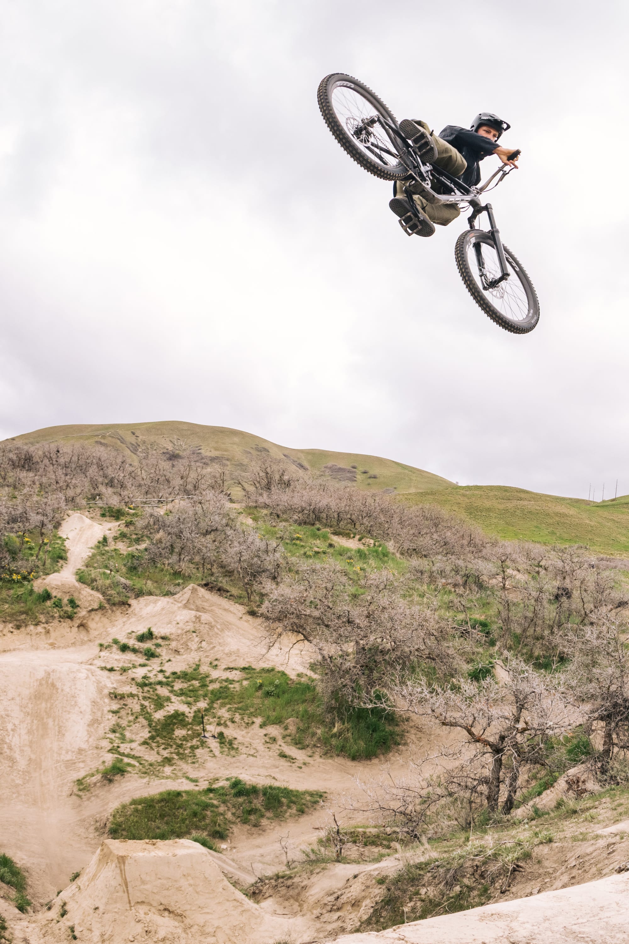 A mountain biker in a helmet and 686 Men's Anything Cargo Pant - Relaxed Fit soars mid-air over a dirt trail, surrounded by grassy hills and leafless trees under a cloudy sky.