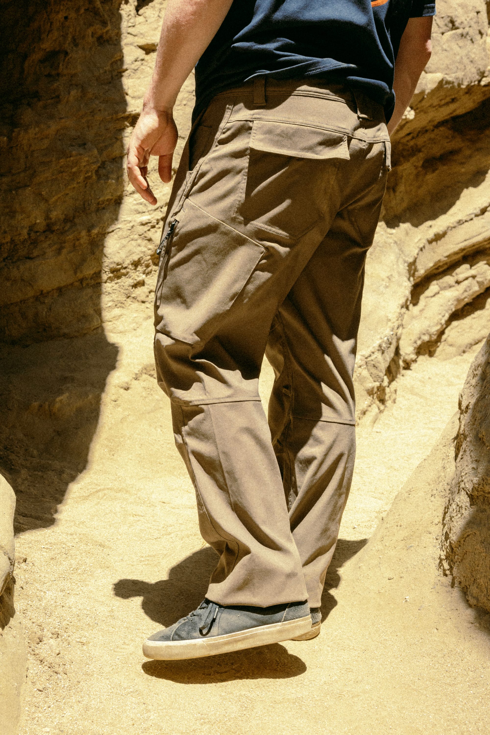 A person wearing 686 Men's Anything Cargo Pant - Relaxed Fit by 686 and black shoes walks up a sandy, rocky path between large rocks in bright sunlight.