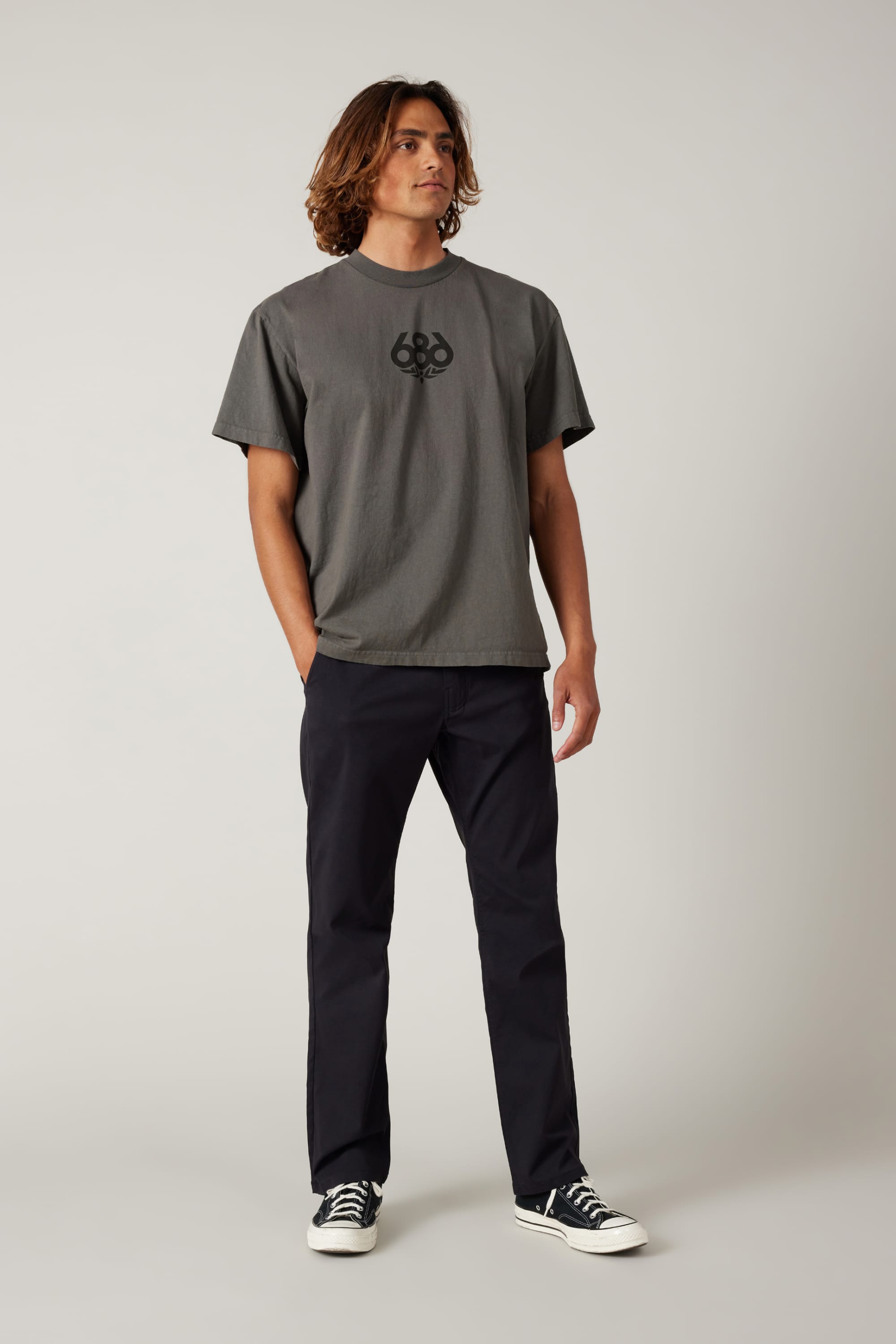 A young man with shoulder-length hair wears a gray t-shirt with a black logo, 686 Men's Everywhere® Pant - Relaxed Fit by 686, and black-and-white sneakers, standing against a plain light background.