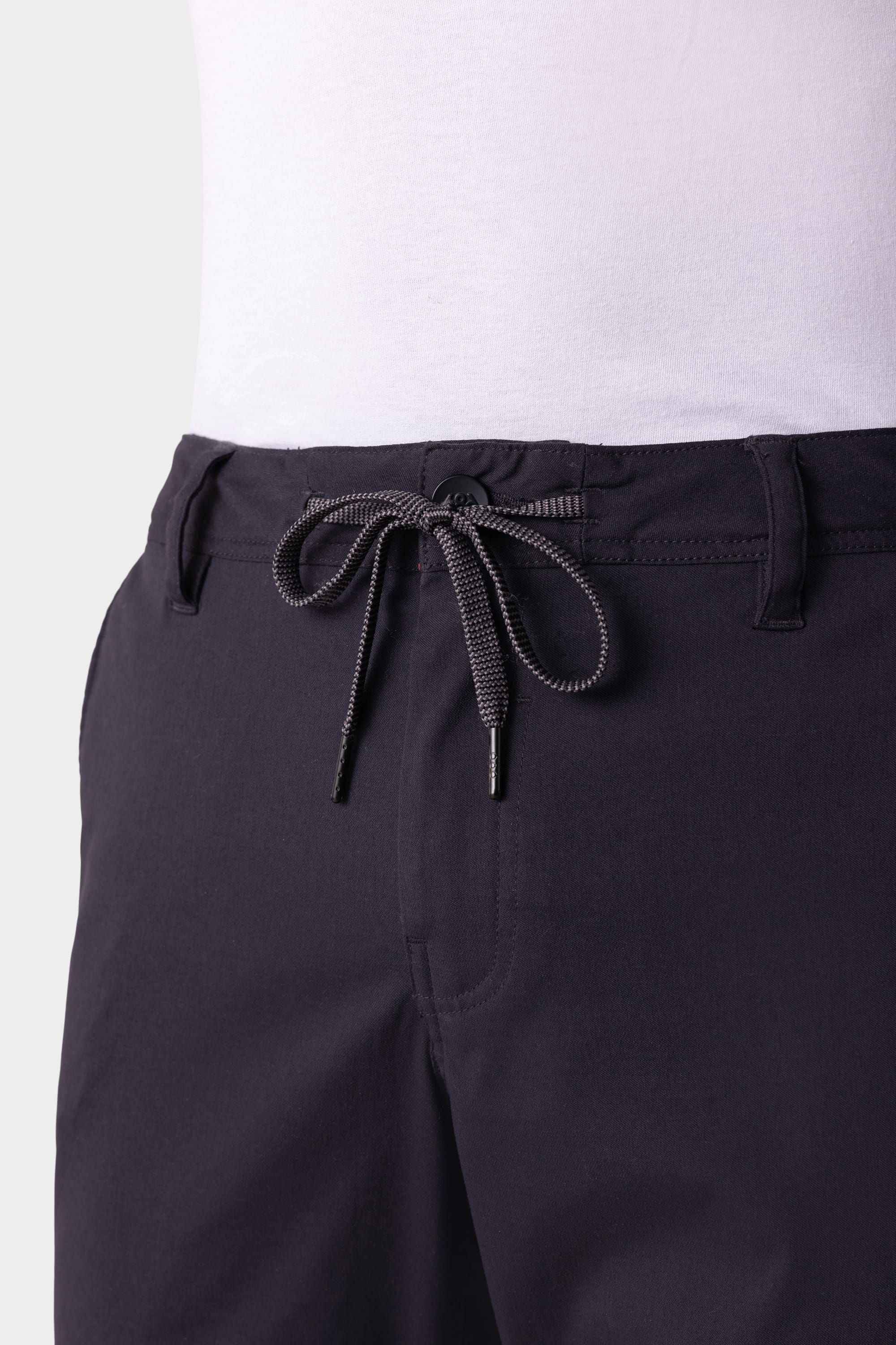 Close-up of a person wearing the 686 Men's Everywhere® Pant - Relaxed Fit in a dark color with a gray drawstring, belt loops, front button closure, and a white shirt tucked in. Brand: 686.