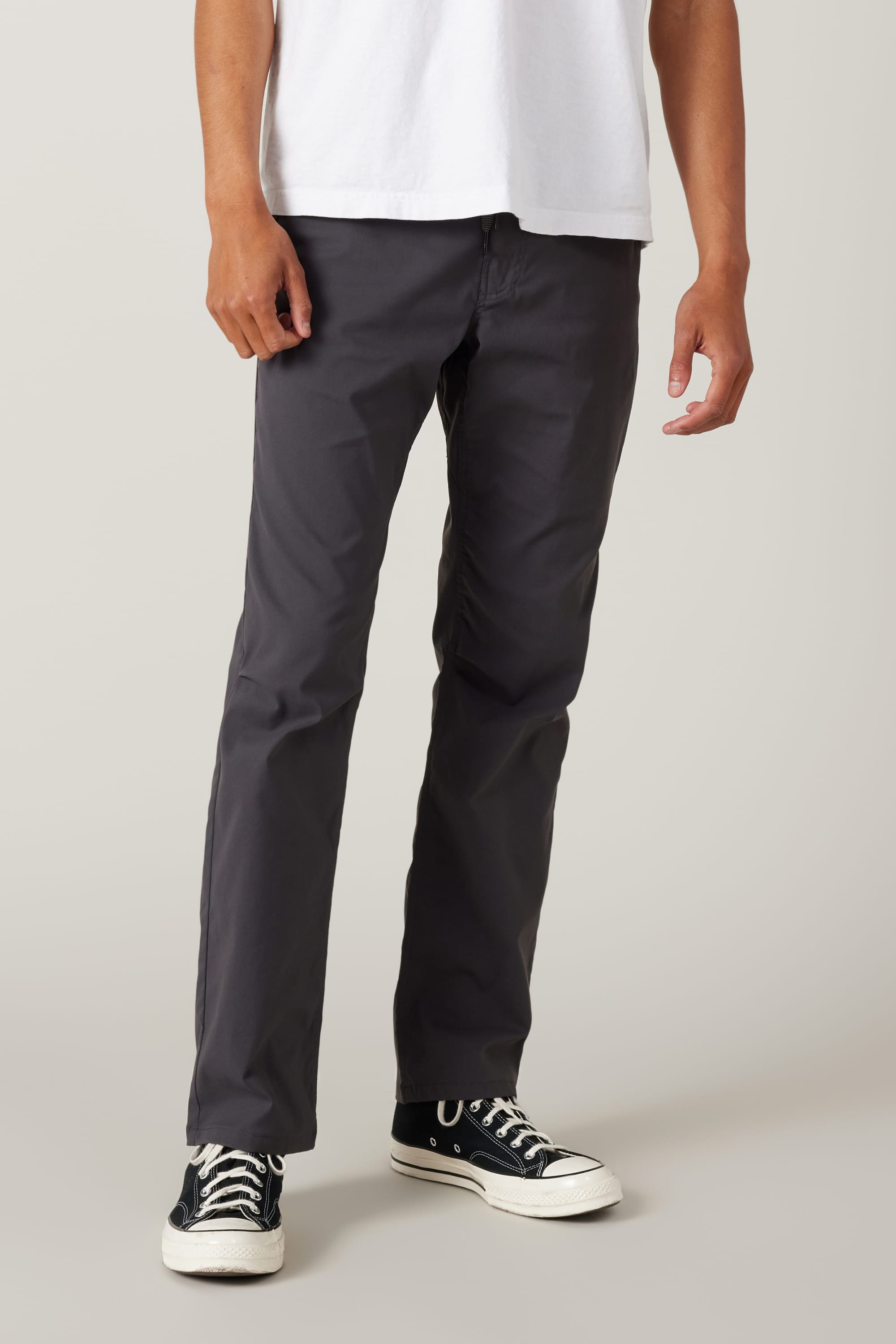 Only the lower half of a person is visible, wearing a white t-shirt, 686 Men's Everywhere® Pant - Relaxed Fit in dark gray, and black Converse sneakers against a light gray background.