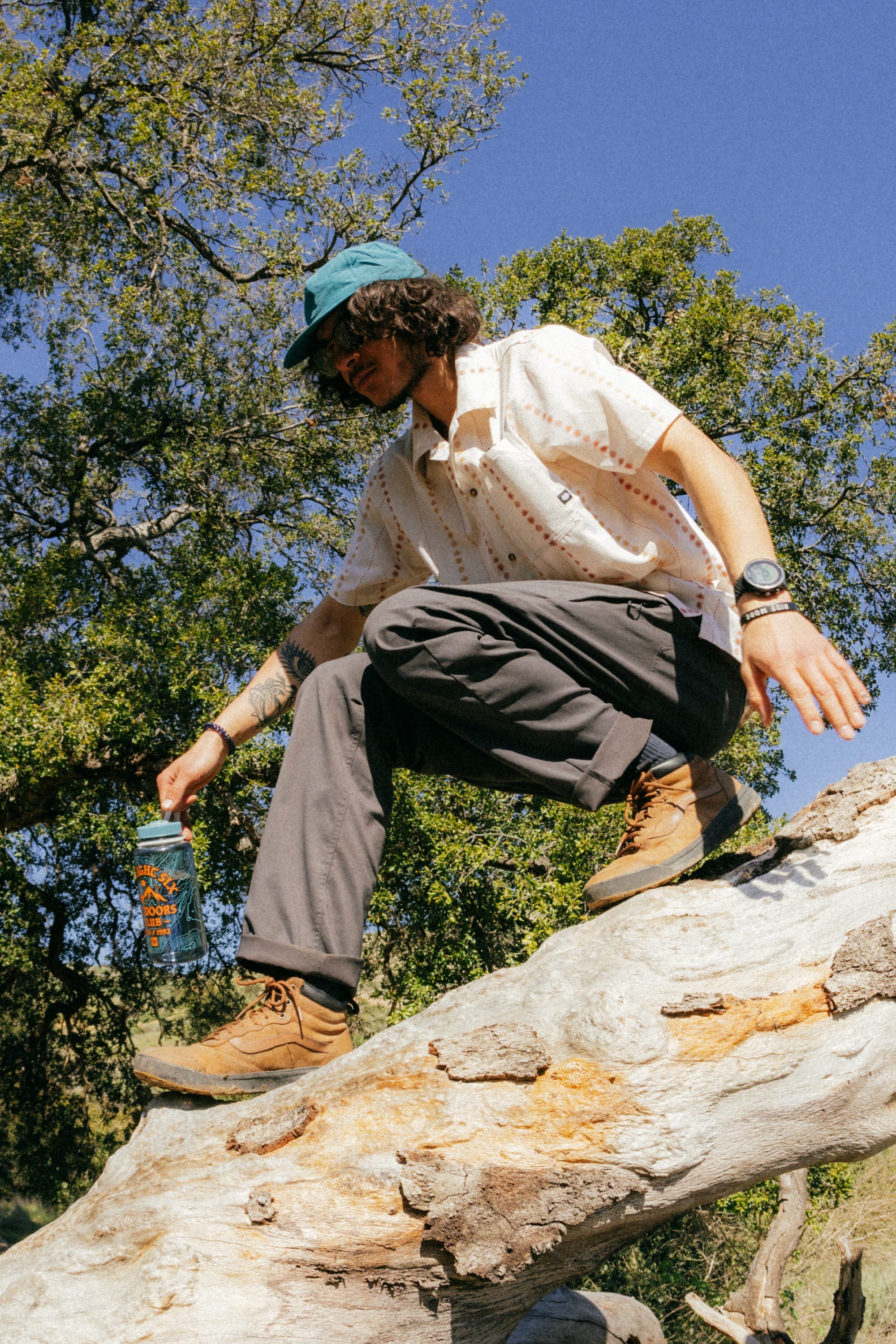 A person wearing 686 Men's Everywhere® Pant - Relaxed Fit by 686, a blue cap, sunglasses, patterned shirt, and hiking boots climbs over a fallen tree trunk in the forest, holding a water bottle amid green trees under a clear sky.