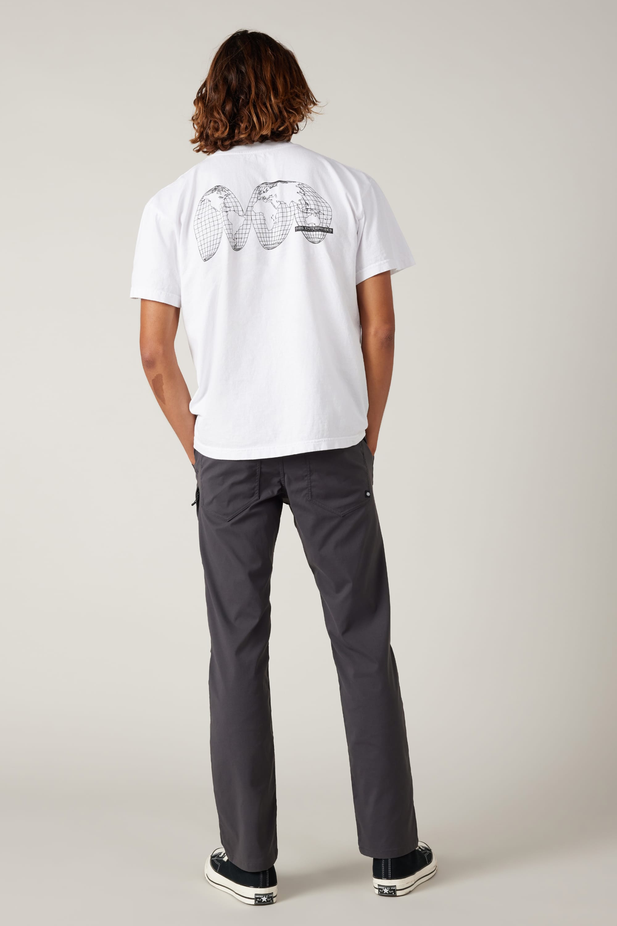 A person with shoulder-length hair stands facing away, wearing a white graphic T-shirt featuring a tennis racket on the back, black sneakers, and 686 Men's Everywhere® Pant - Relaxed Fit by 686. The background is plain light gray.