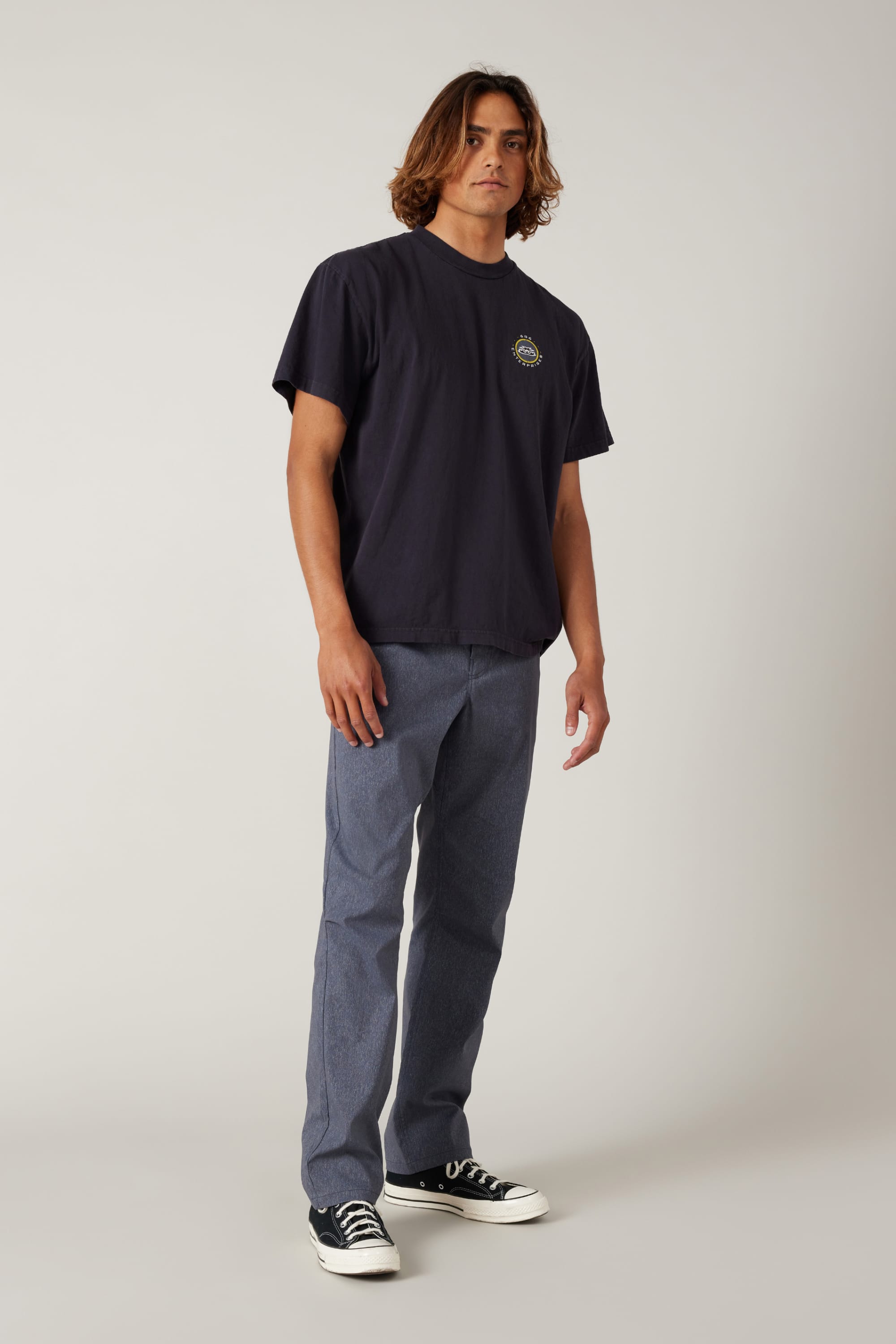 A young man with wavy hair wears a loose black t-shirt, 686 Men's Everywhere® Pant - Relaxed Fit by 686 in blue, and black-and-white sneakers while standing against a plain light background.