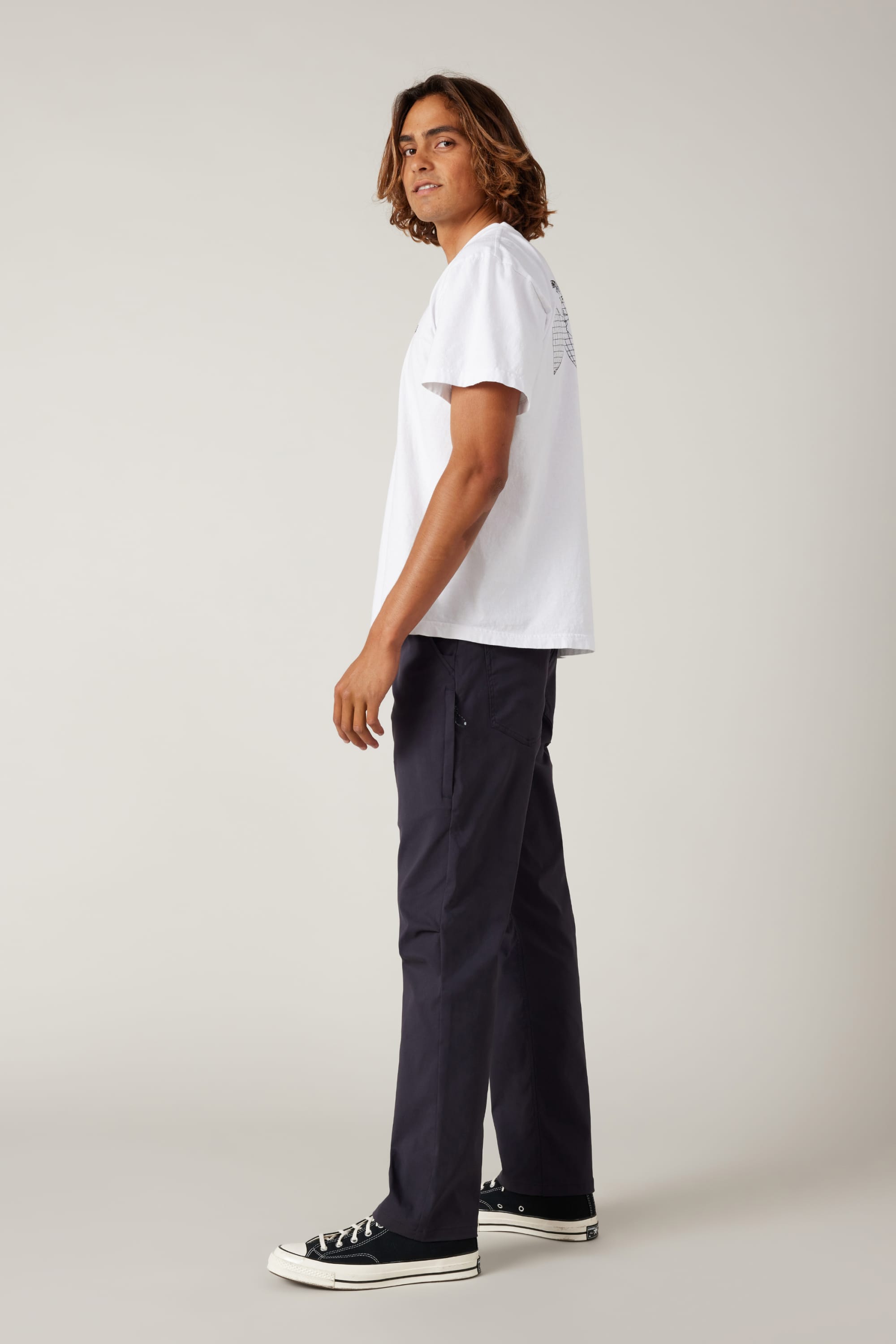A person with shoulder-length hair stands sideways, wearing a white T-shirt, 686 Men's Everywhere® Pant - Relaxed Fit by 686, and black sneakers, looking over their shoulder against a plain light background.