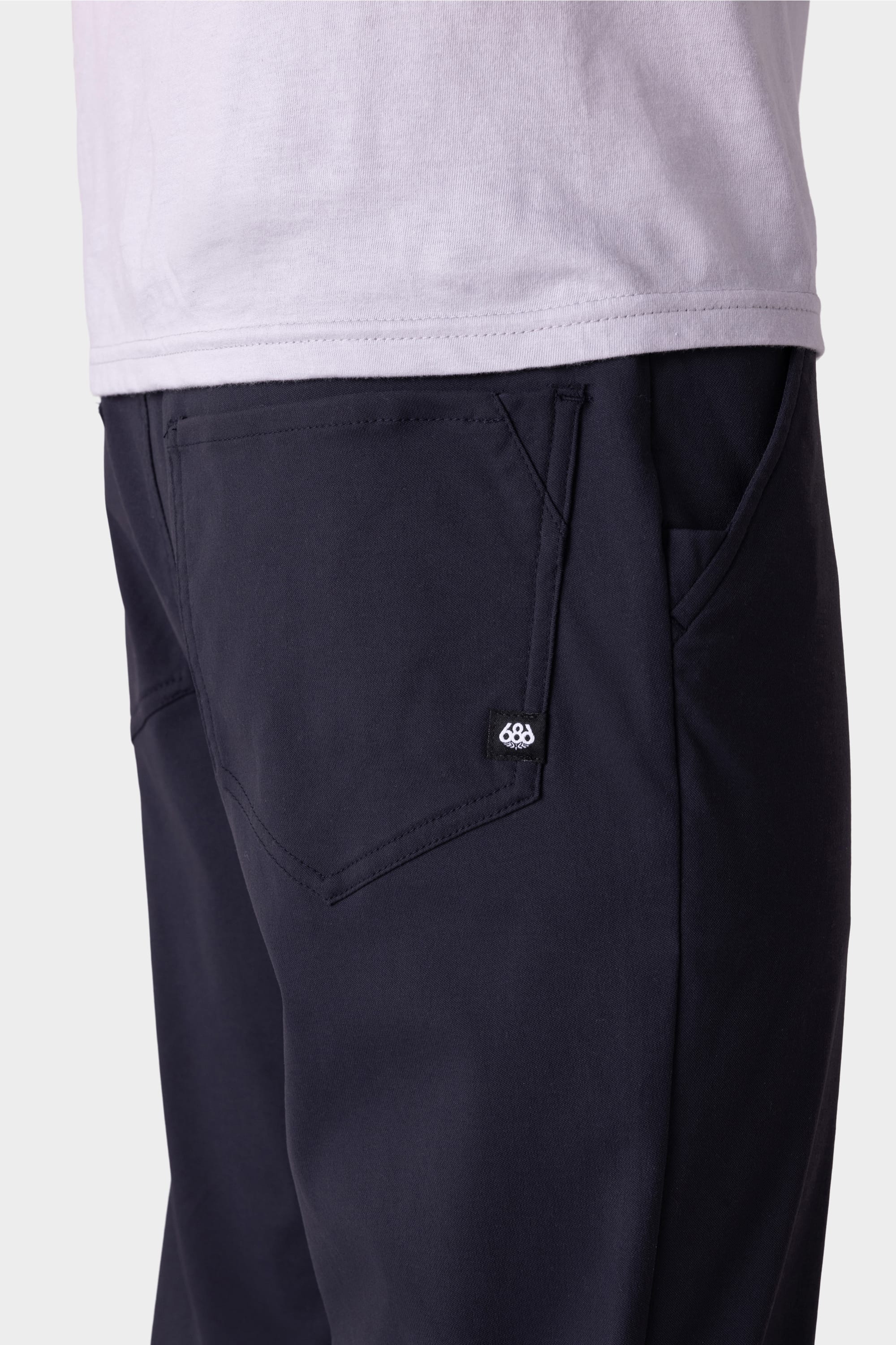 Close-up of a person wearing the 686 Men's Everywhere® Pant - Relaxed Fit in dark navy with a light shirt. Focus is on the left side pocket, showcasing a small black tag with the white 686 logo.