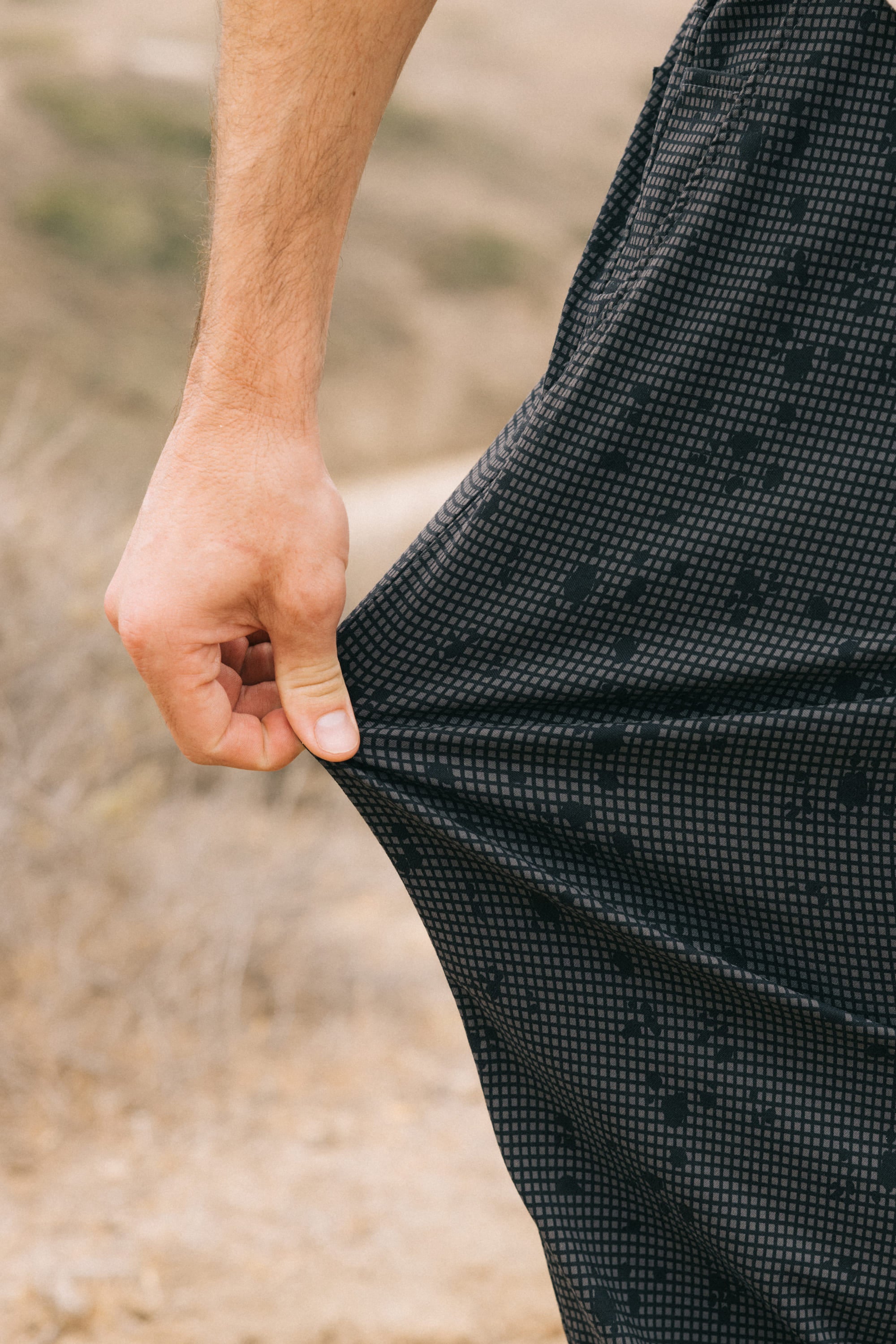 A person's hand stretches the fabric of 686 Men's Everywhere® Pant - Relaxed Fit by 686, showcasing its breathable, black patterned material outdoors against a blurred natural background.