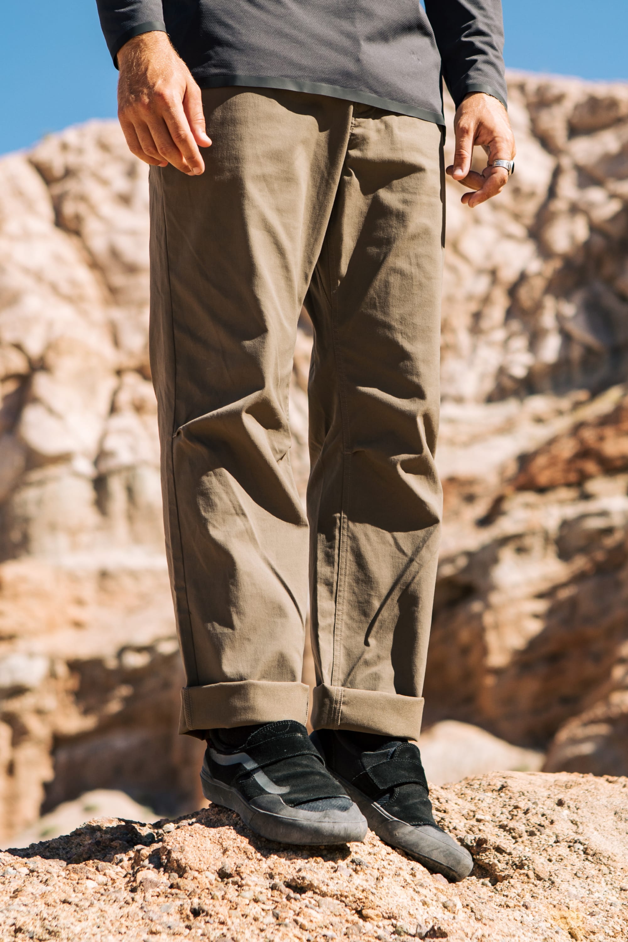 A person wearing 686 Men's Everywhere® Pant - Relaxed Fit by 686 and black sneakers stands on rocky terrain with blurred rock formations in the background.
