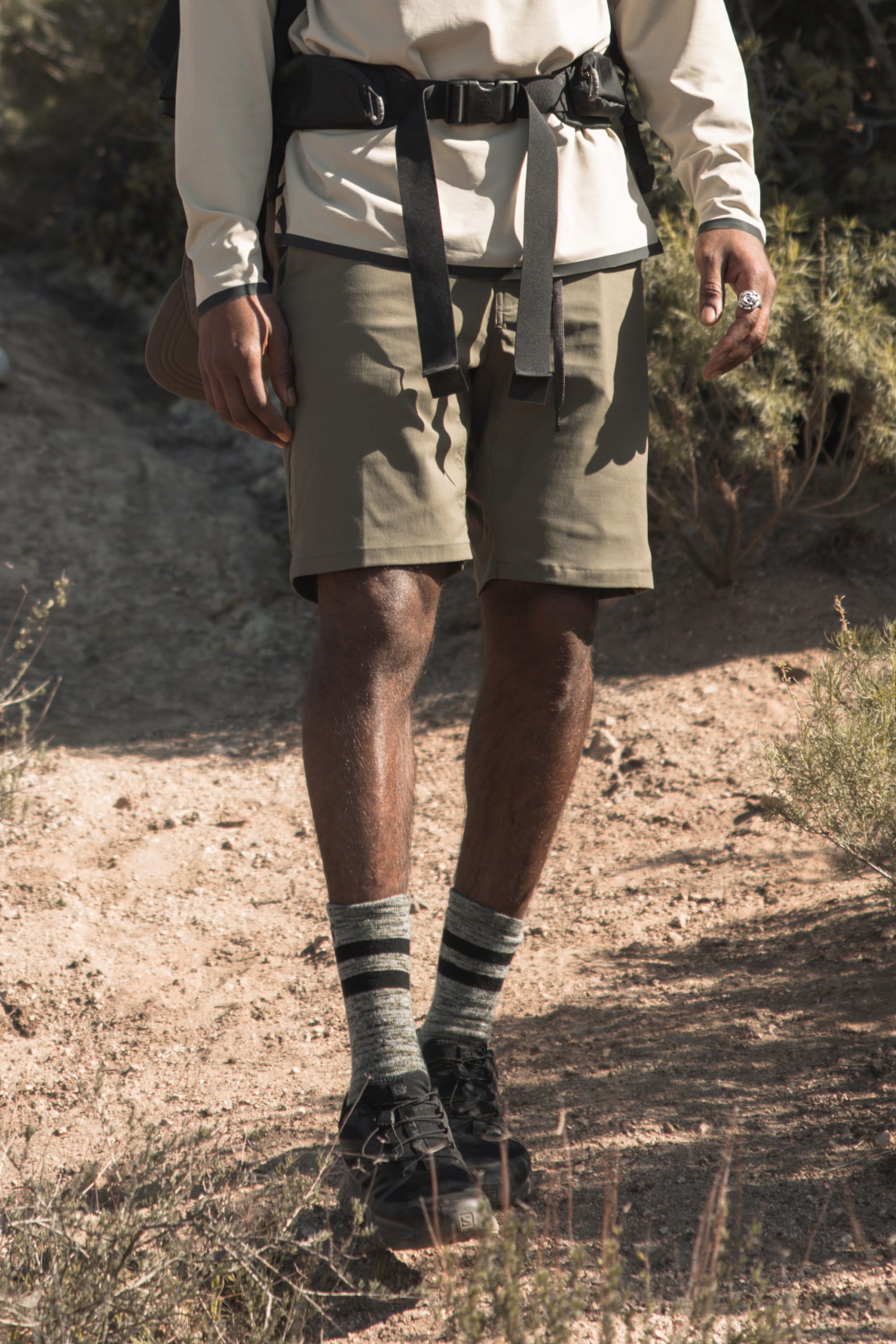 Wearing a long-sleeve shirt, 686 Everywhere® Hybrid Short by 686, striped socks, and hiking boots, a person with a backpack walks on a sunlit dirt trail amid dry plants.