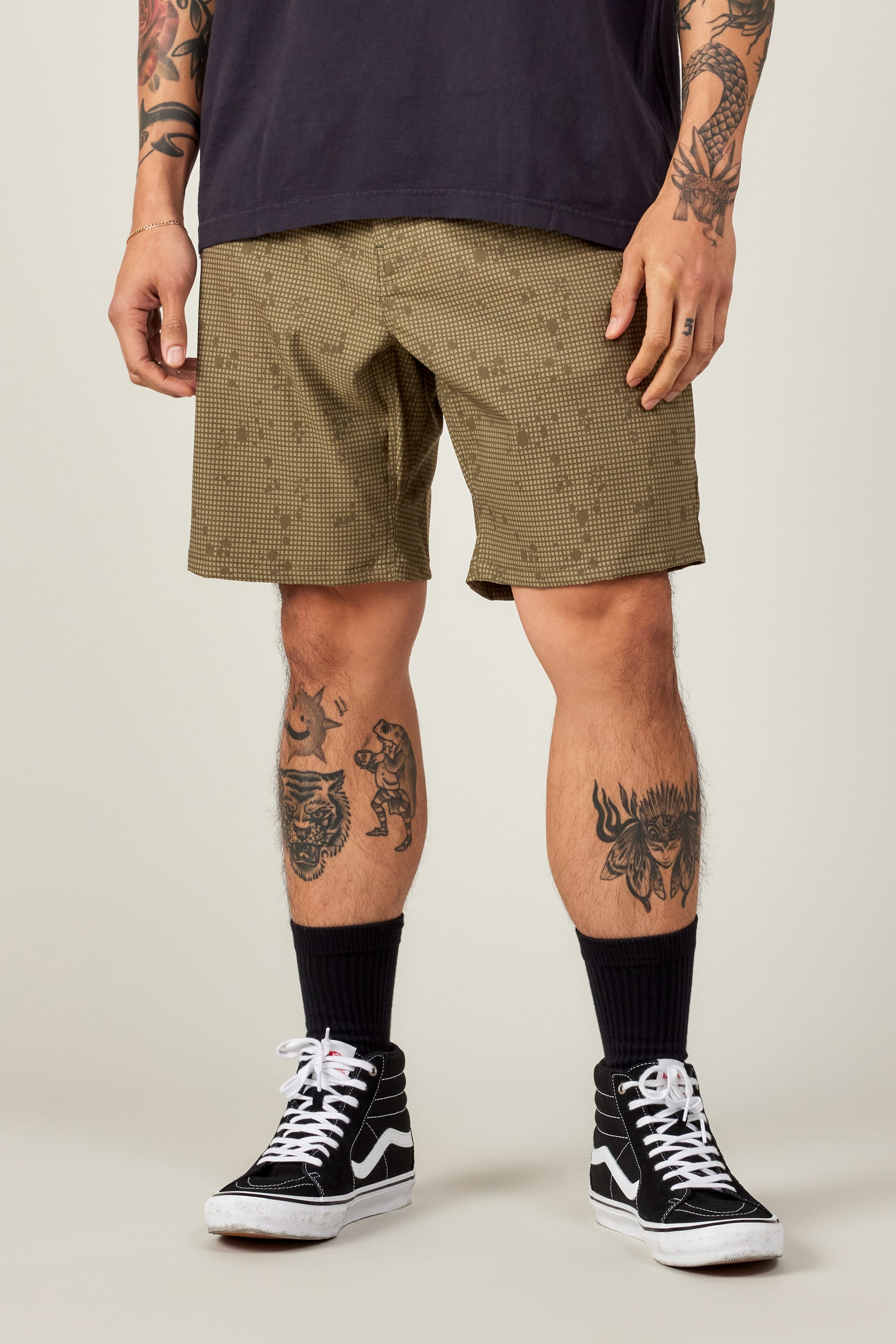 A person wearing a black T-shirt, 686 Everywhere® Hybrid Short by 686 in brown checkered print, black socks, and black Vans sneakers. Their tattooed arms and legs stand out against a plain light background.