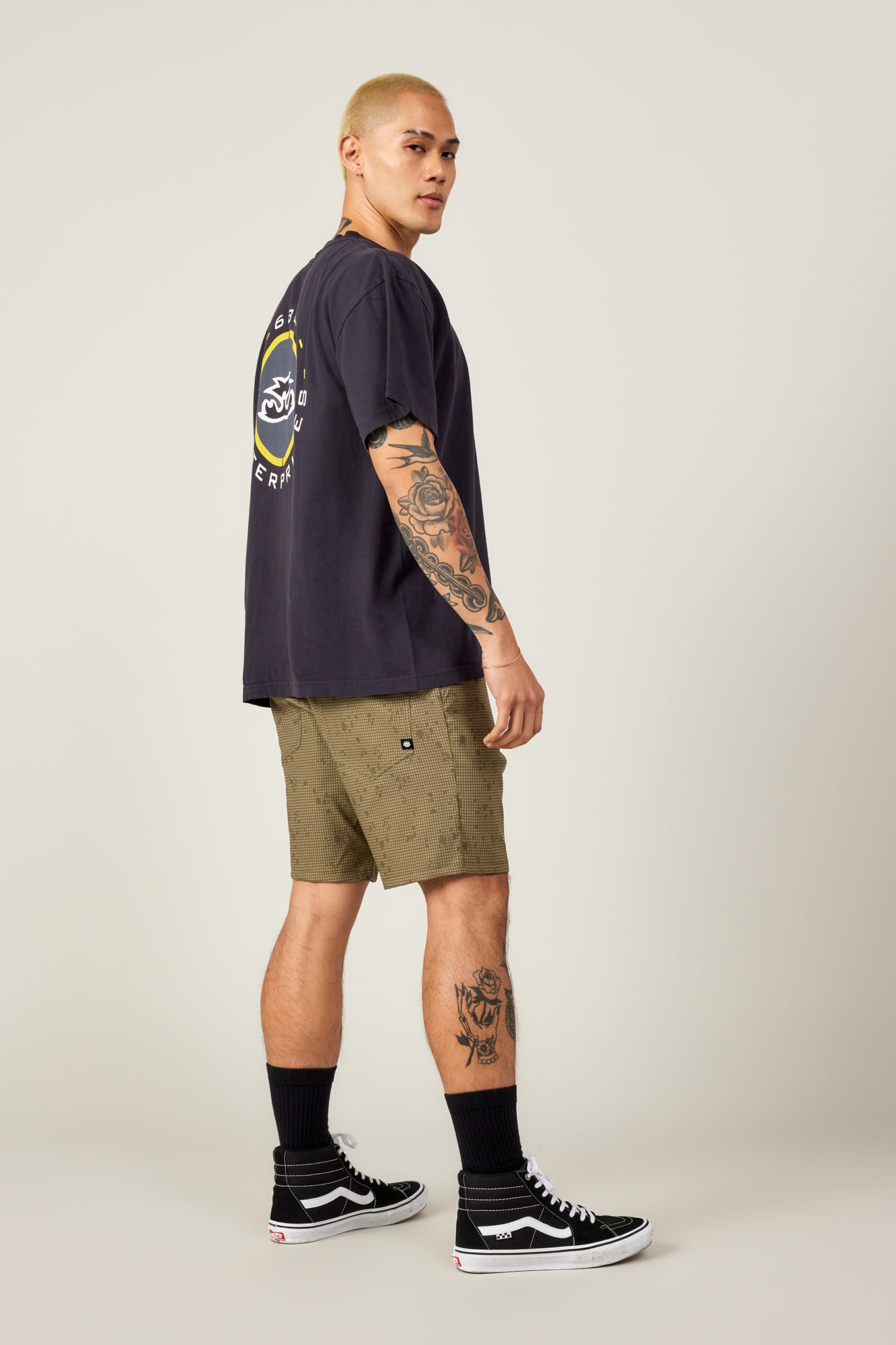 A person with short blond hair and tattoos stands in a side pose, wearing a black tee with a round back graphic, 686 Everywhere® Hybrid Short by 686, black crew socks, and black Vans sneakers.