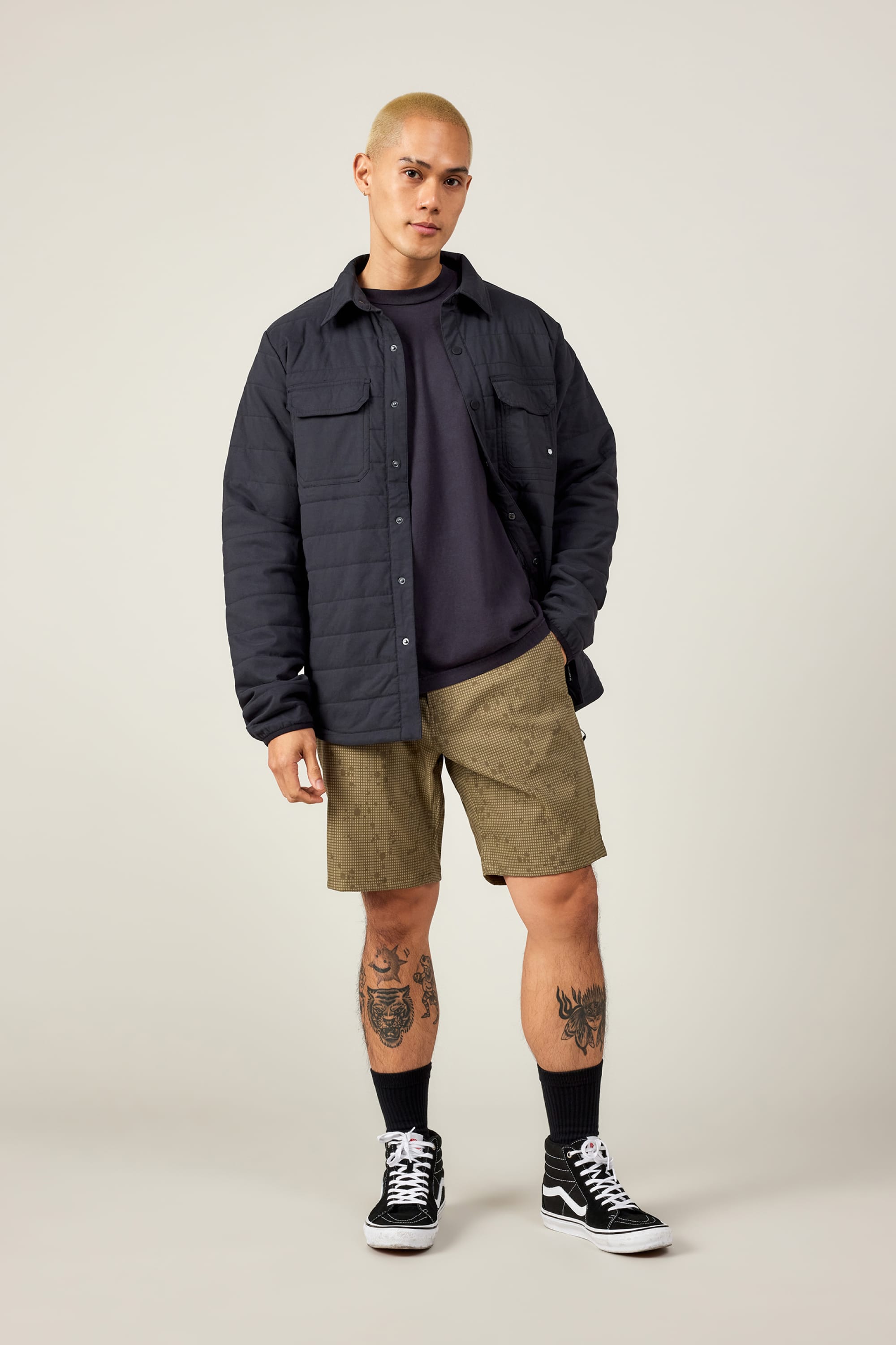 A person with short blond hair wears a black quilted jacket, dark shirt, black socks, black sneakers, and 686 Everywhere® Hybrid Short by 686 in olive green, standing against a plain background and showing leg tattoos.