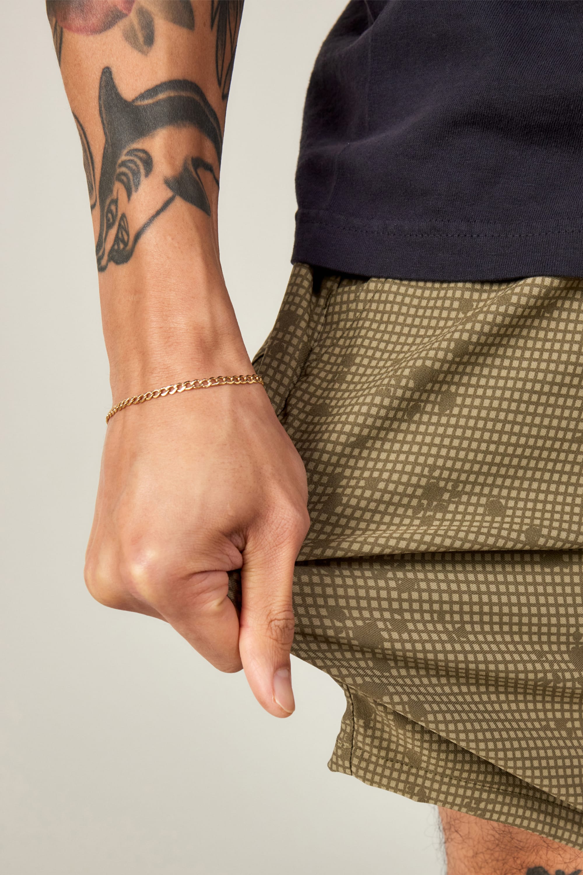 A close-up shows a tattooed forearm with a gold chain bracelet pulling the hem of 686 Everywhere® Hybrid Short, highlighting the green patterned, stain-resistant design and stylish practicality from 686.