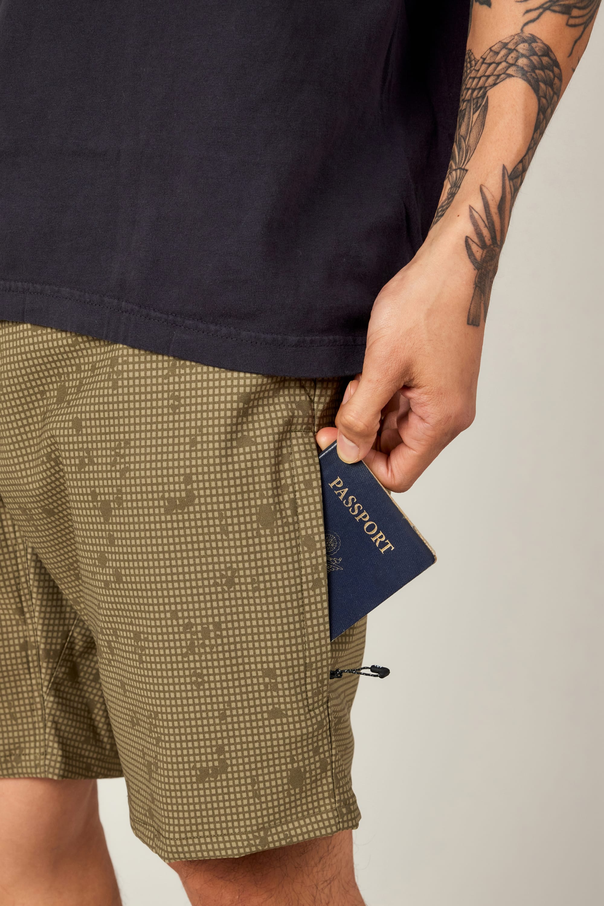 A person in a black shirt and patterned green 686 Everywhere® Hybrid Short holds a blue passport partly tucked in their pocket, with a visible tattoo on their arm.