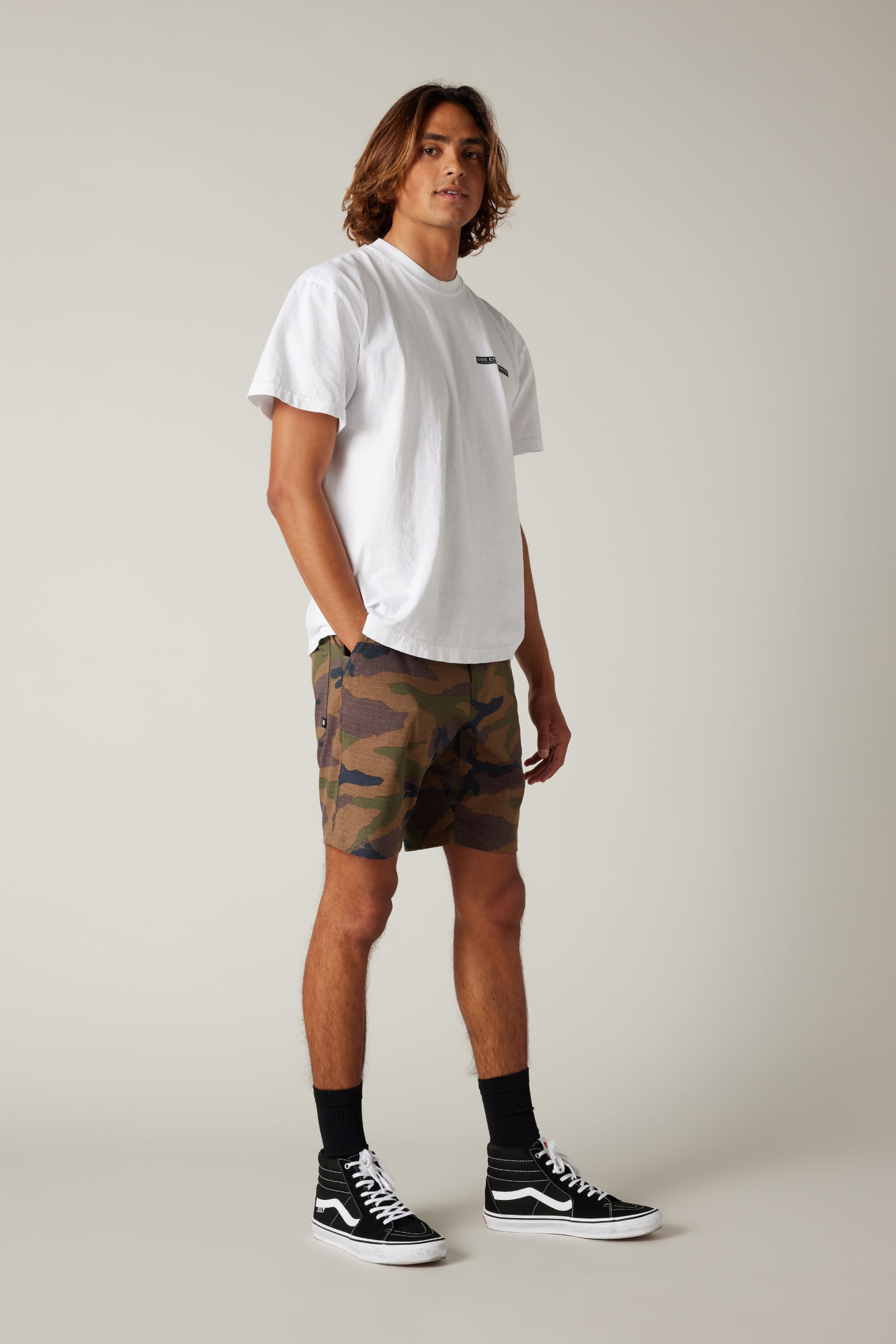 A person with medium-length brown hair stands against a plain background, wearing a white t-shirt and 686 Everywhere® Hybrid Short, paired with black socks and black Vans sneakers, with hands tucked in the shorts’ pockets.