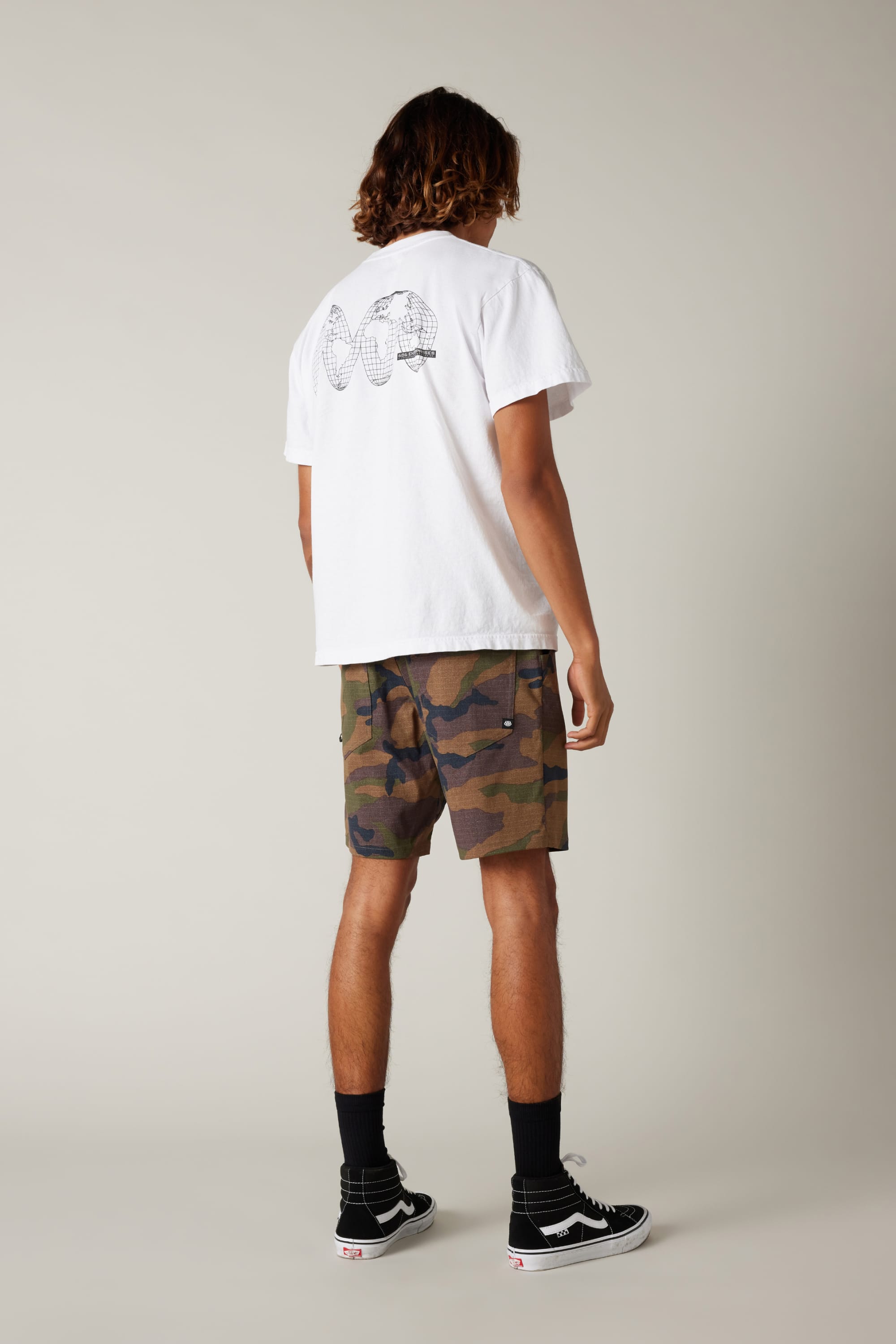 A person stands facing away wearing a white T-shirt with a black geometric graphic, 686 Everywhere® Hybrid Short camo shorts from 686, black socks, and black Vans sneakers against a plain light background.