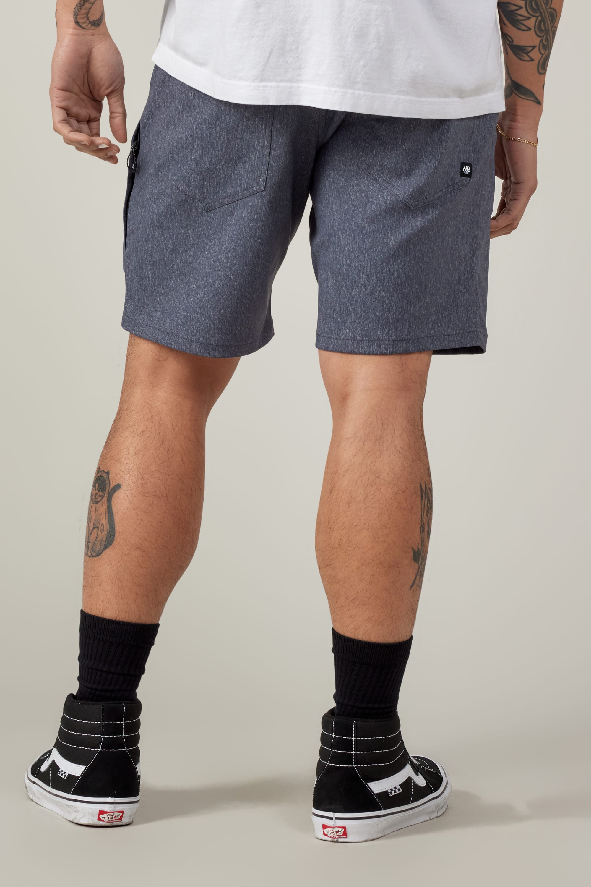 A person in a white T-shirt, 686 Everywhere® Hybrid Short by 686, black socks, and black Vans sneakers stands with their back to the camera, showing leg tattoos.