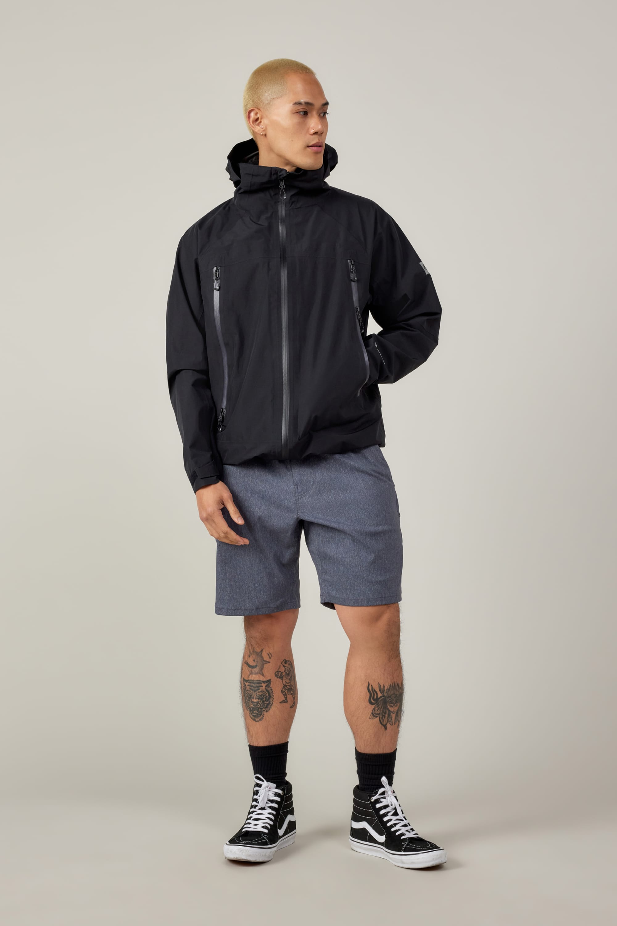 A person with short blond hair wears a black hooded jacket, 686 Everywhere® Hybrid Shorts by 686, black socks, and black-and-white sneakers. They have leg tattoos and stand against a plain light background, looking to the side.