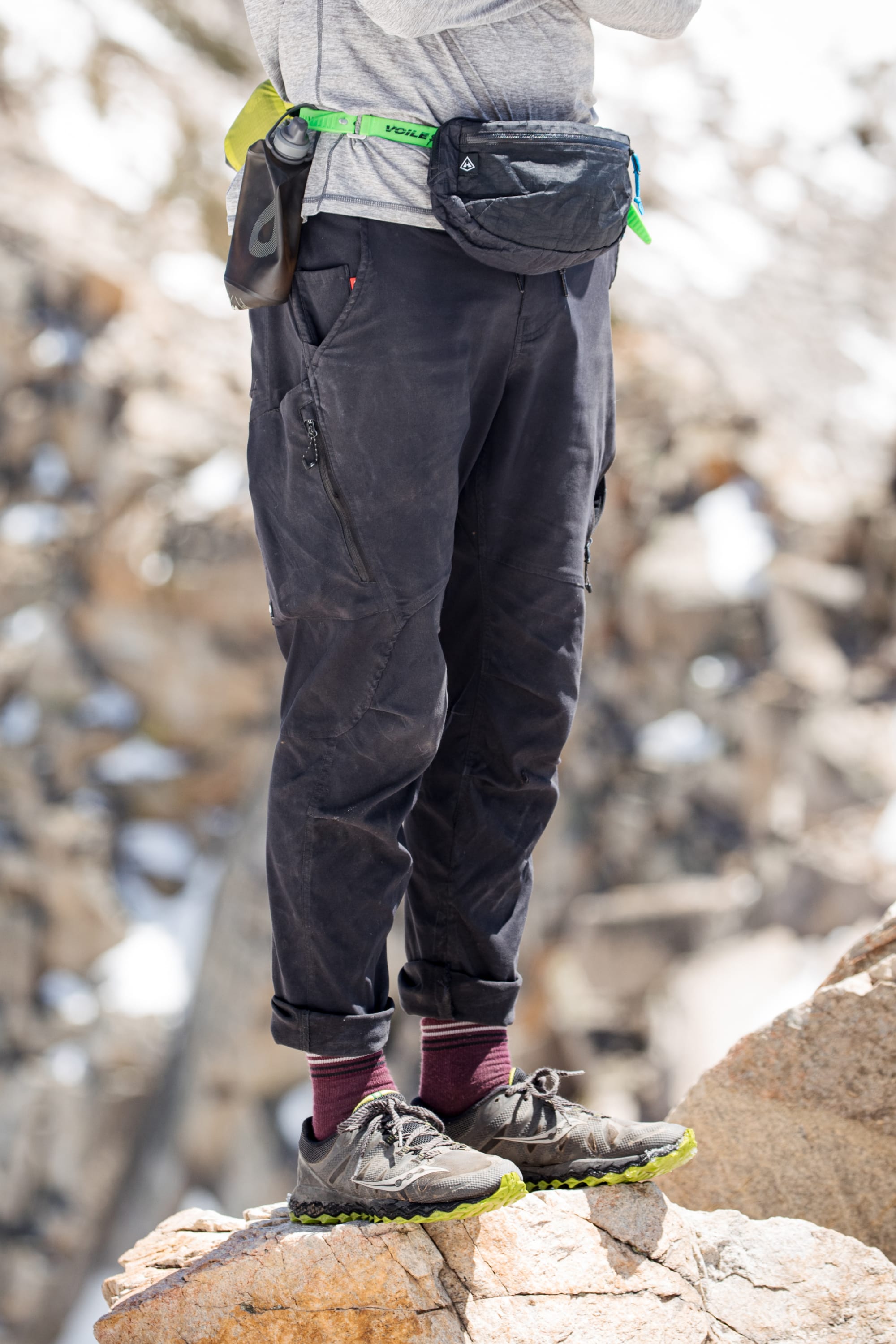 A person stands on rocky terrain, sporting the 686 Men's Anything Cargo Pant - Slim Fit, paired with black trail shoes (green soles), maroon socks, a gray shirt, black waist pack, and bright green belt. Their upper body is not visible, ready for adventure.