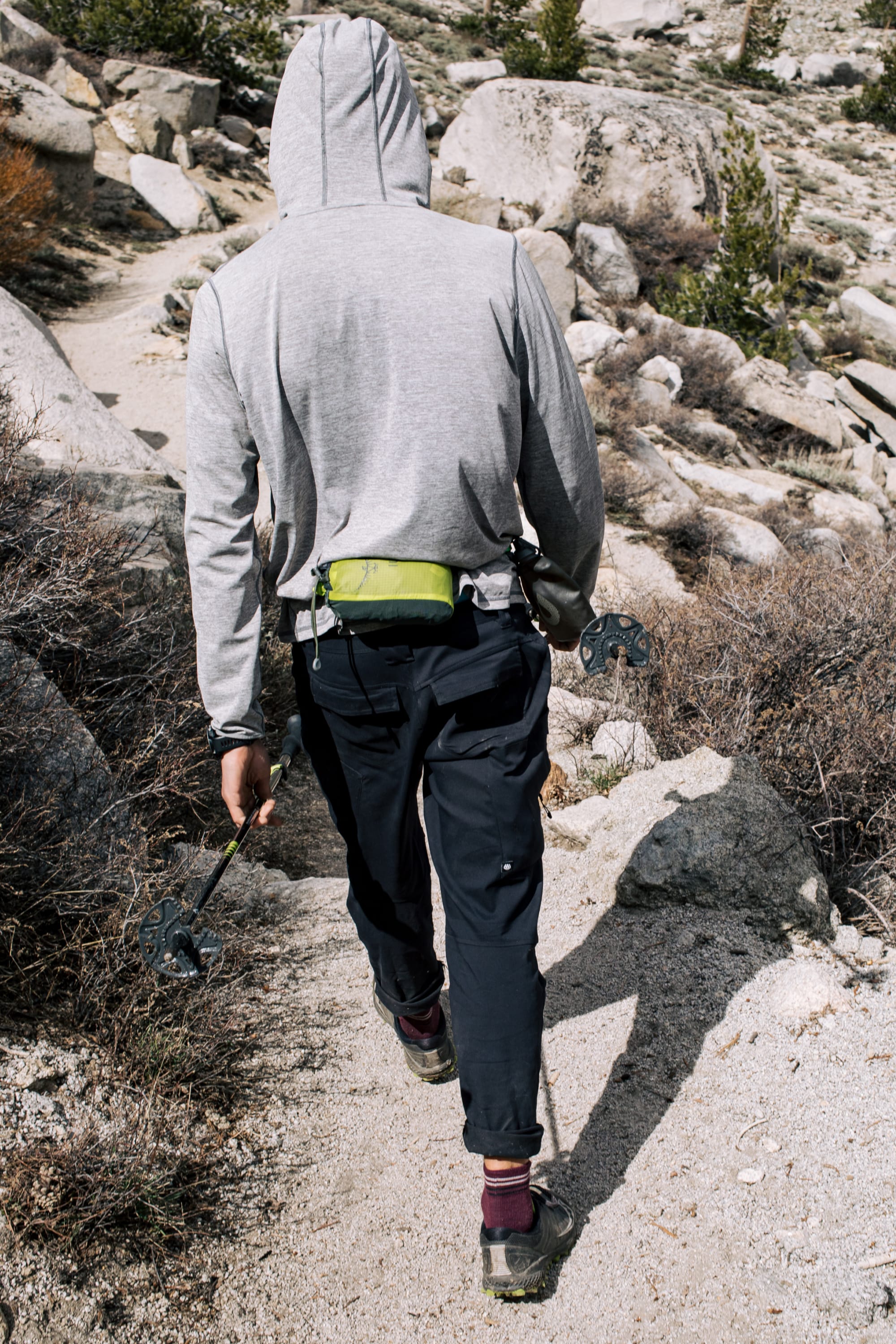 Wearing the 686 Men's Anything Cargo Pant - Slim Fit, a hiker tackles a rocky trail with trekking poles and a small green bag, ready for adventure among shrubs and large rocks.