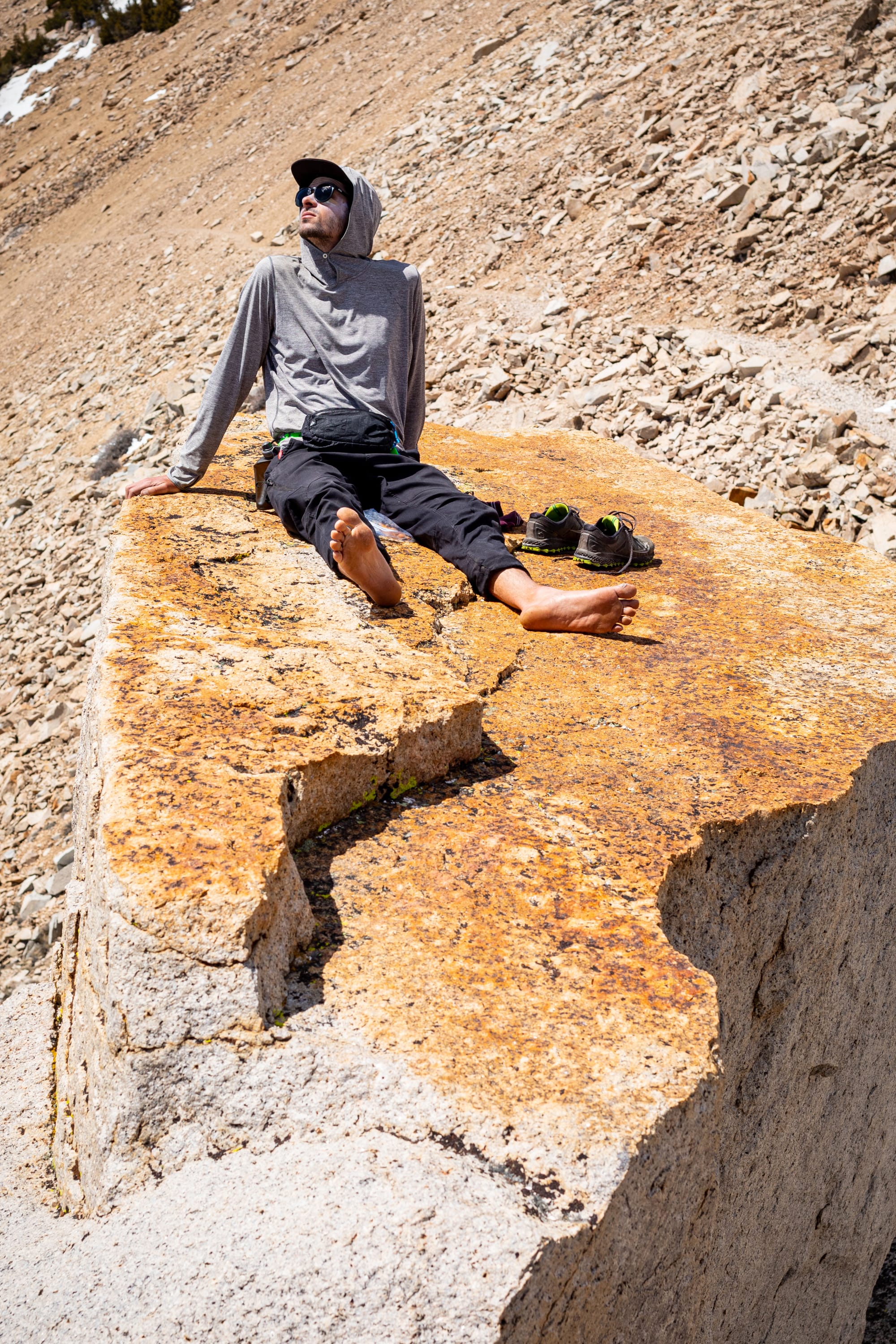 A person in a hoodie and sunglasses relaxes barefoot on a mountain rock, their 686 Men's Anything Cargo Pant - Slim Fit by 686 and shoes placed beside them, soaking up the sun in the rugged landscape.