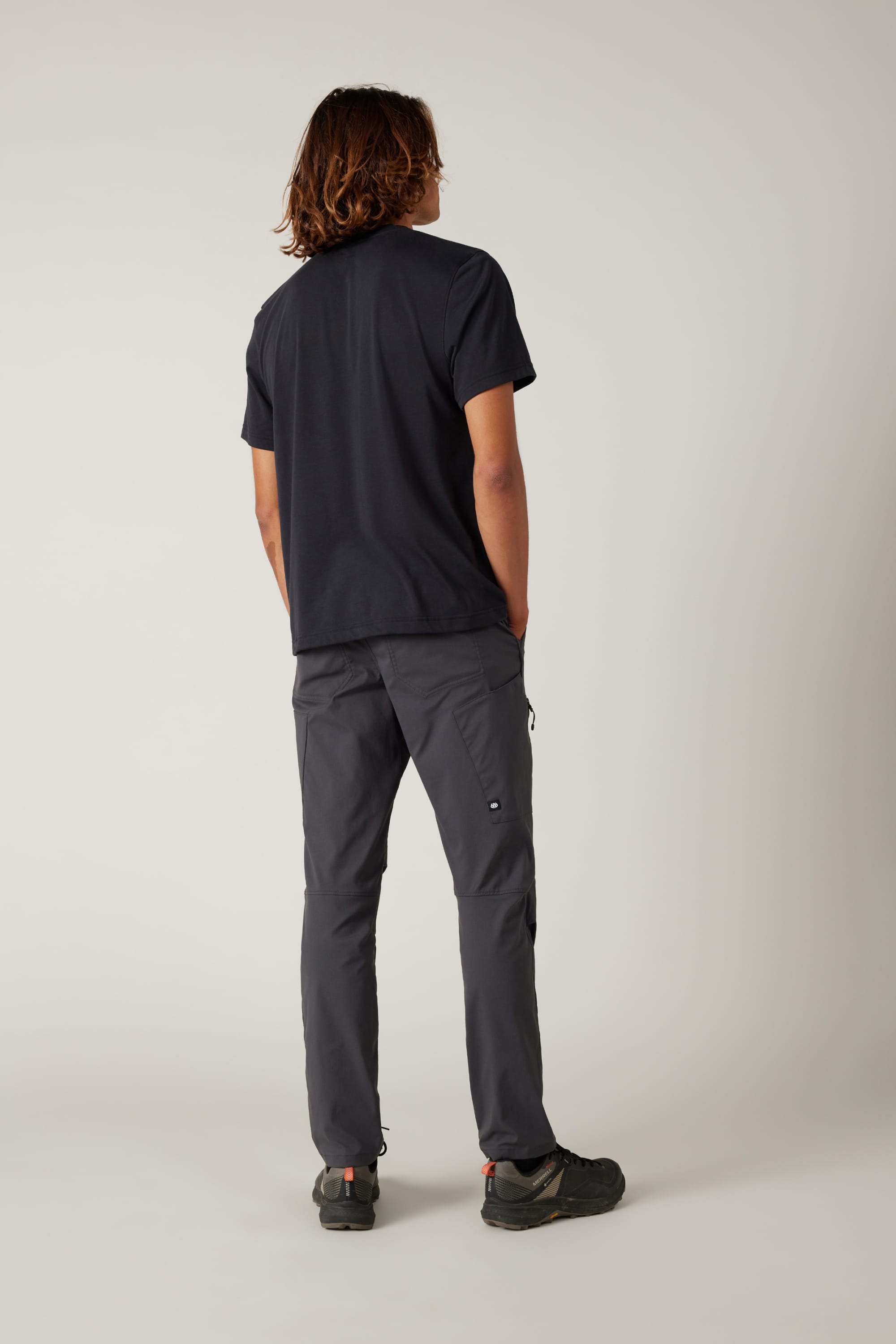 A person with shoulder-length hair stands facing away in a black t-shirt, 686 Men's Anything Cargo Pant - Slim Fit by 686, and black shoes against a plain light background—ready for any adventure.