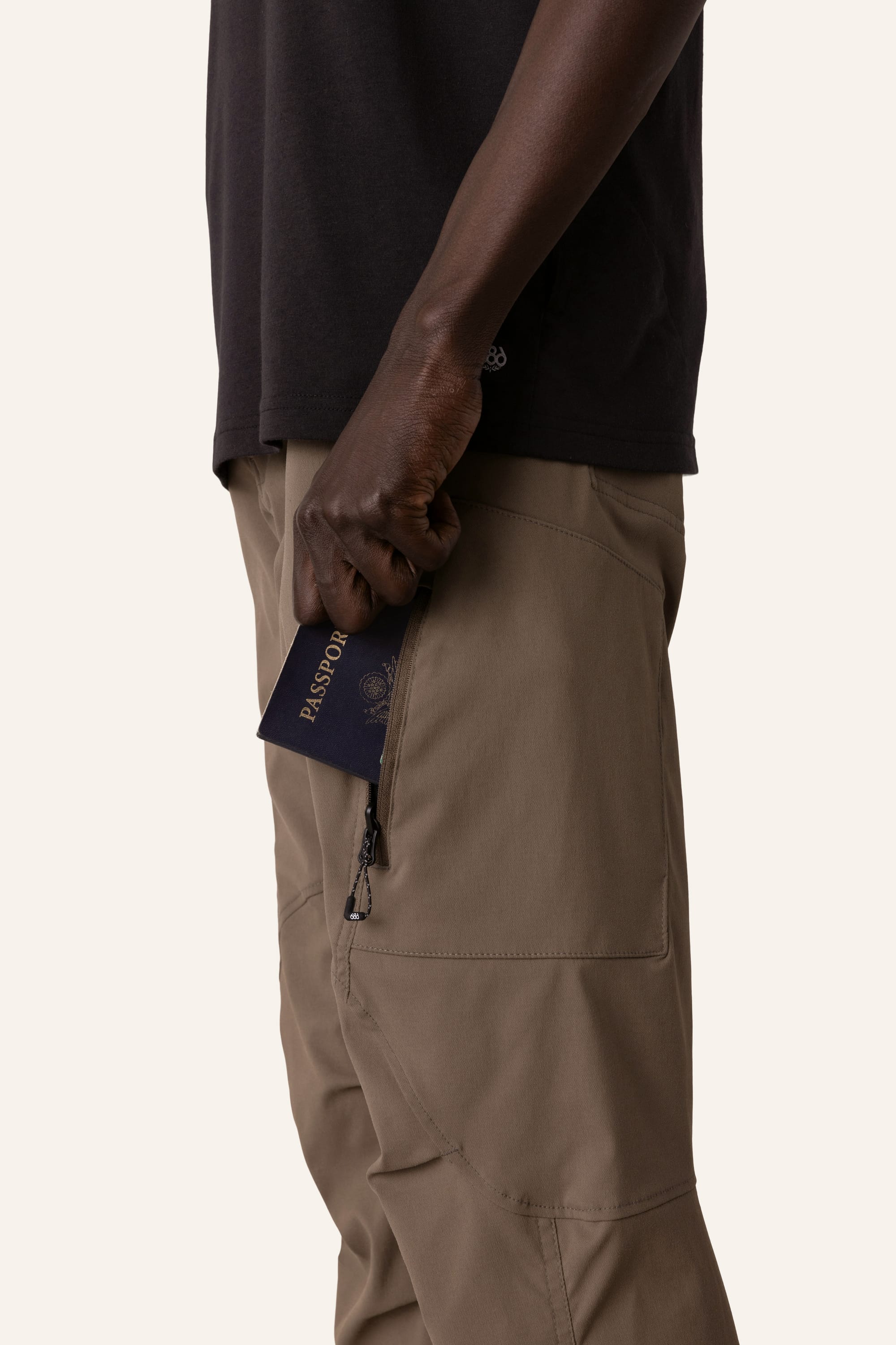 A person in a black shirt tucks a navy passport into the zippered side pocket of their 686 Men's Anything Cargo Pant - Slim Fit by 686. The plain light background hints at adventure ahead.