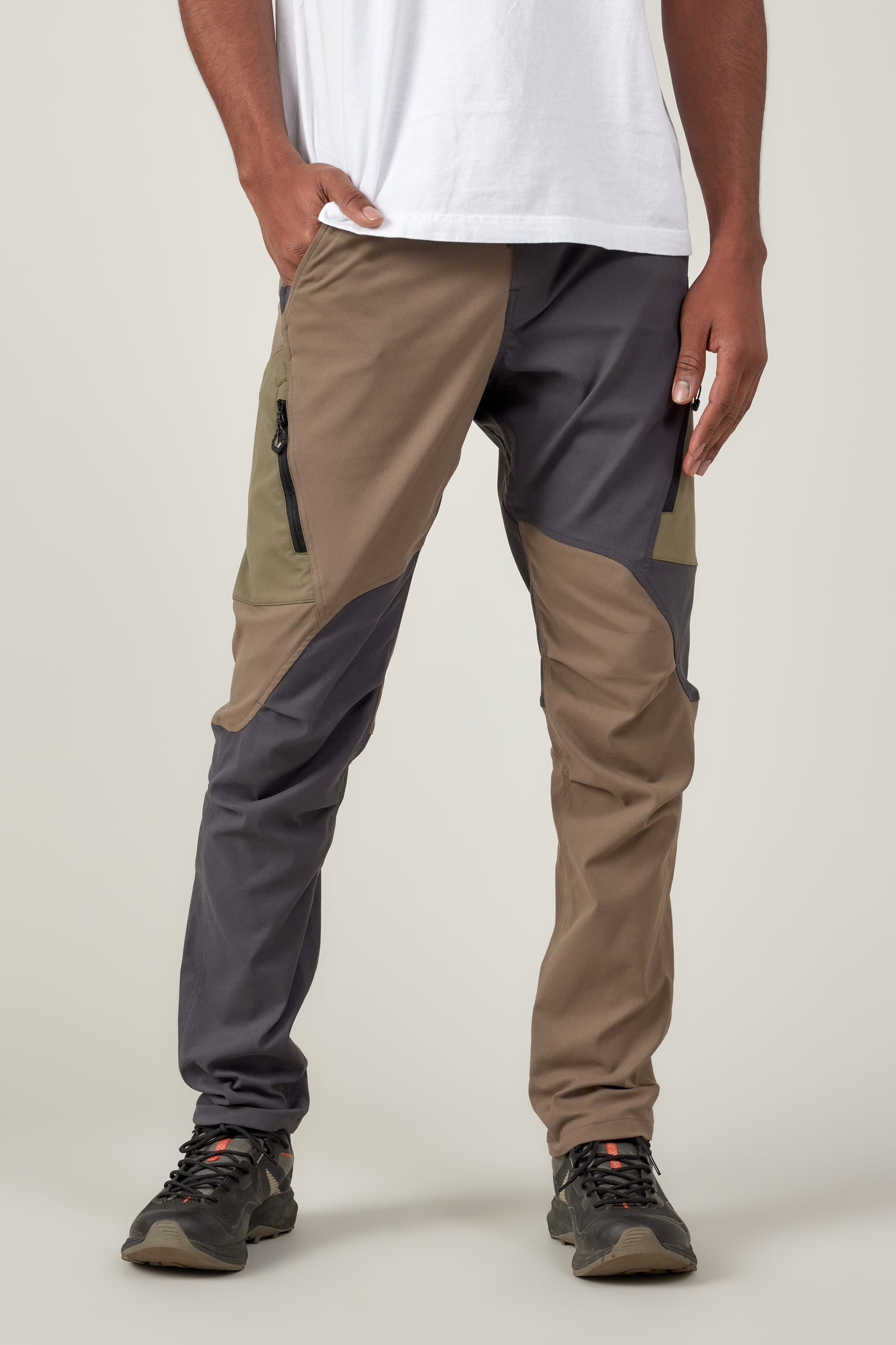 A person in 686 Men's Anything Cargo Pant - Slim Fit by 686, featuring color-blocked brown, olive, and gray panels, a white T-shirt, and hiking shoes stands with one hand in their pocket against a plain light background.