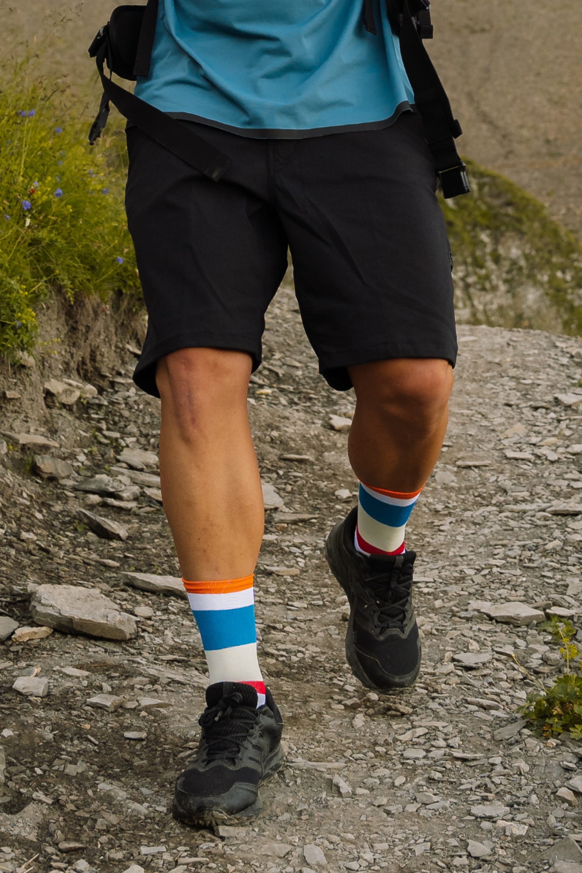 A person wearing 686 Men's Everywhere® Hybrid Short - Relaxed Fit by 686, a blue shirt, colorful striped socks, and black hiking shoes walks on a rocky outdoor trail, with only their legs and lower torso visible.