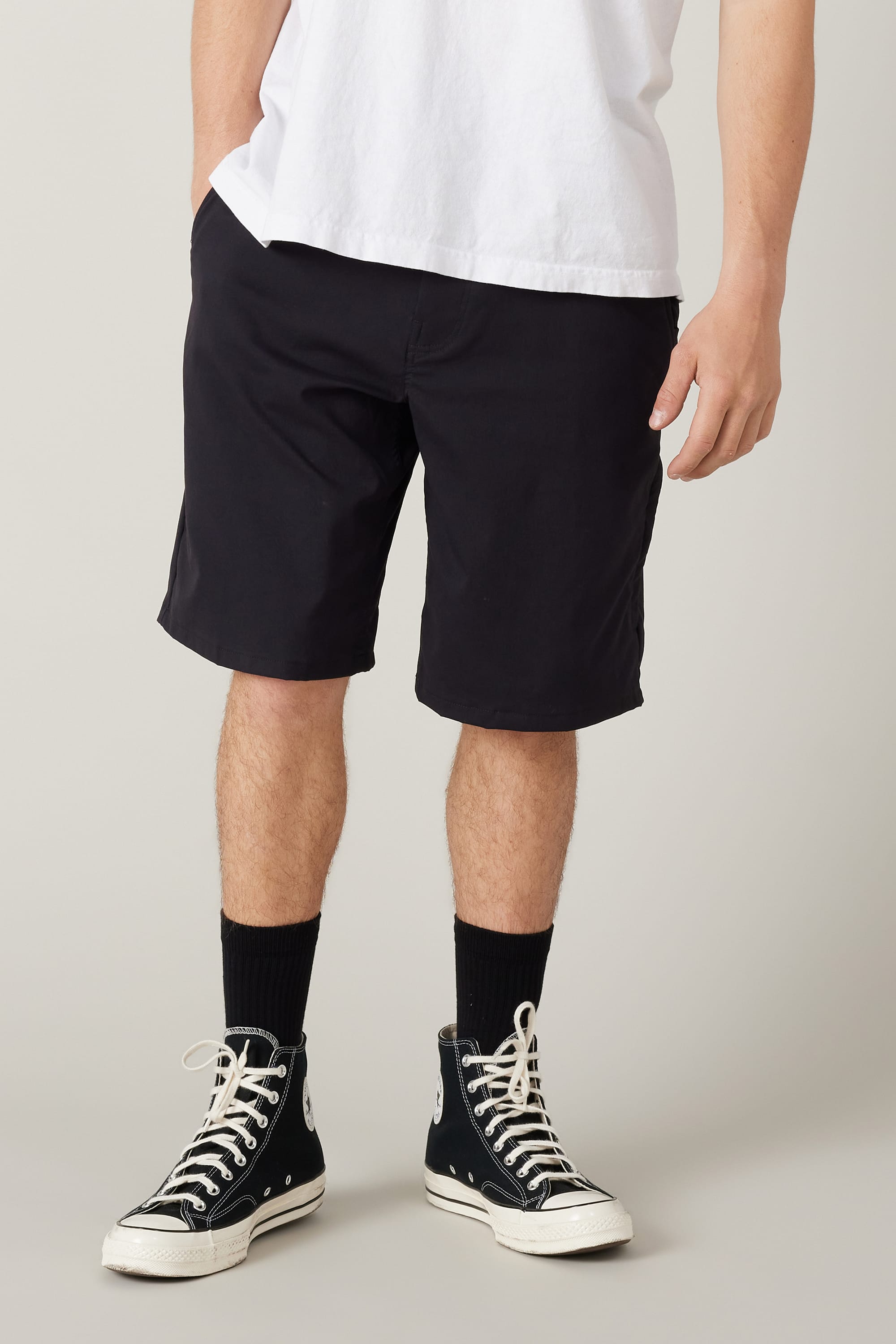 A person wears a white T-shirt and 686 Men's Everywhere® Hybrid Short - Relaxed Fit in black, paired with black crew socks and high-top sneakers, standing against a plain light background. Photo shows from shoulders down.