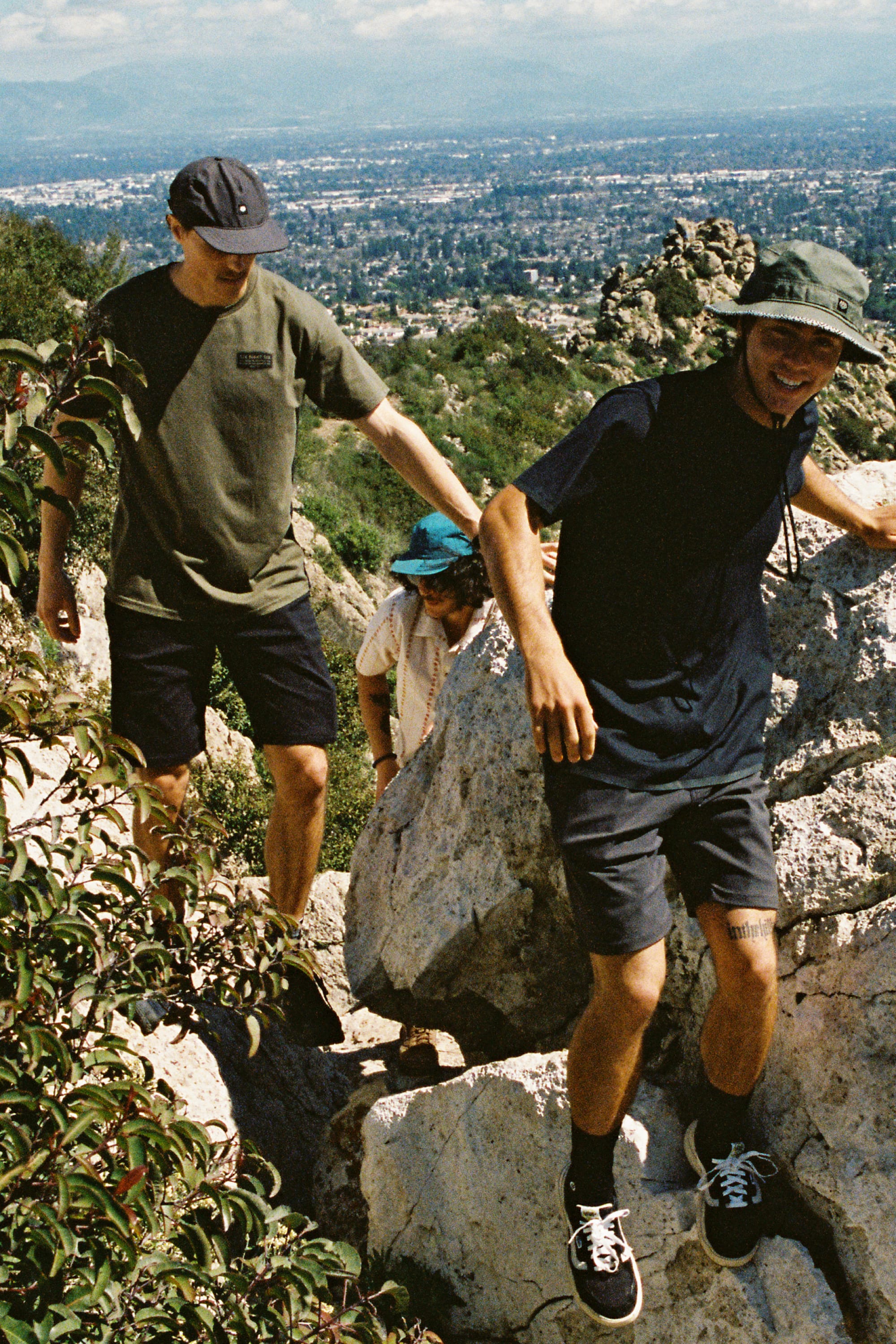 Three people in casual clothes and hats, featuring the 686 Men's Everywhere® Hybrid Short - Relaxed Fit, hike up rocky terrain surrounded by greenery, with a distant cityscape visible on a sunny day.