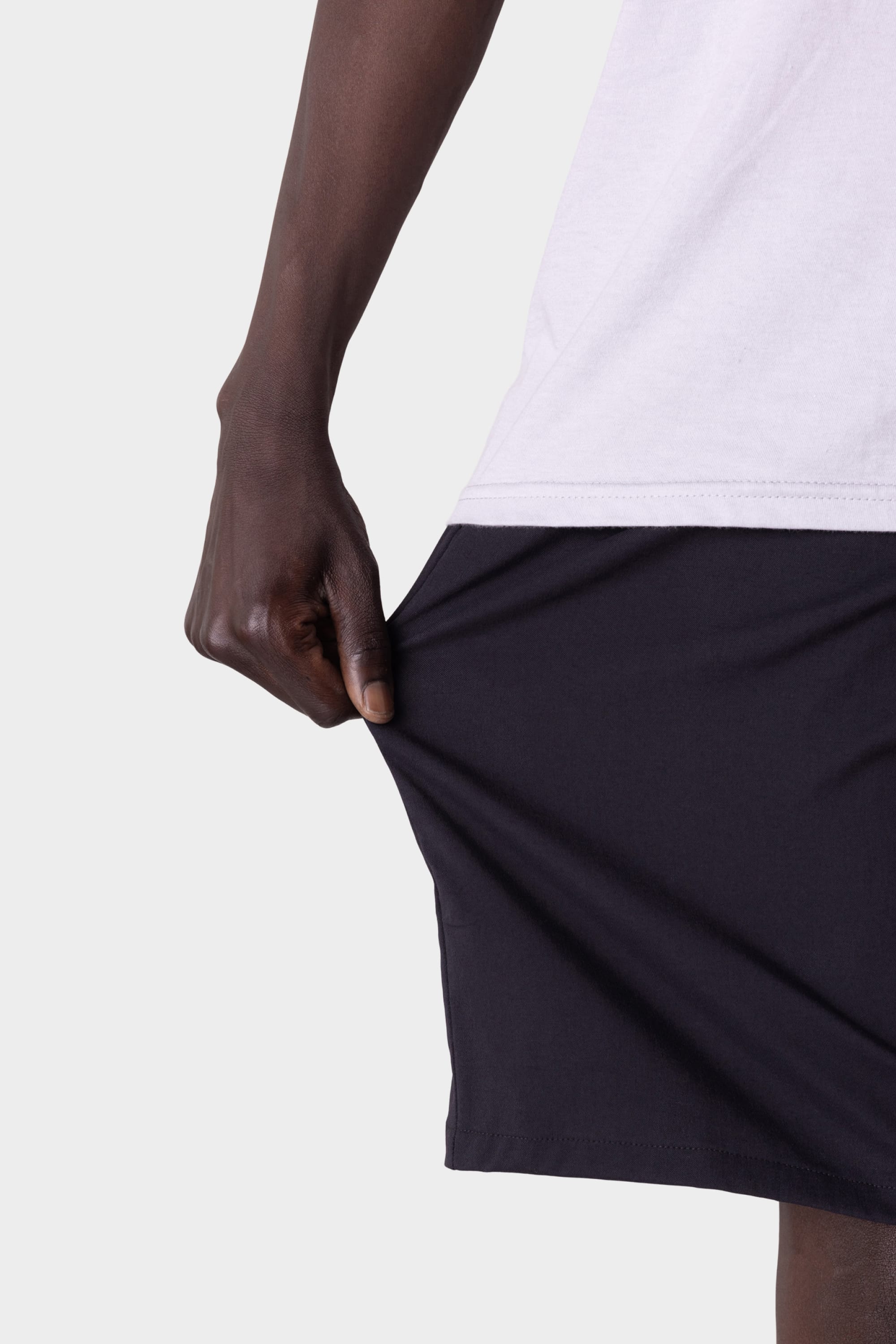 A person wearing a white shirt and 686 Men's Everywhere® Hybrid Short - Relaxed Fit in black pulls at the shorts' hem with one hand, highlighting the stretchy fabric by 686 against a plain background.
