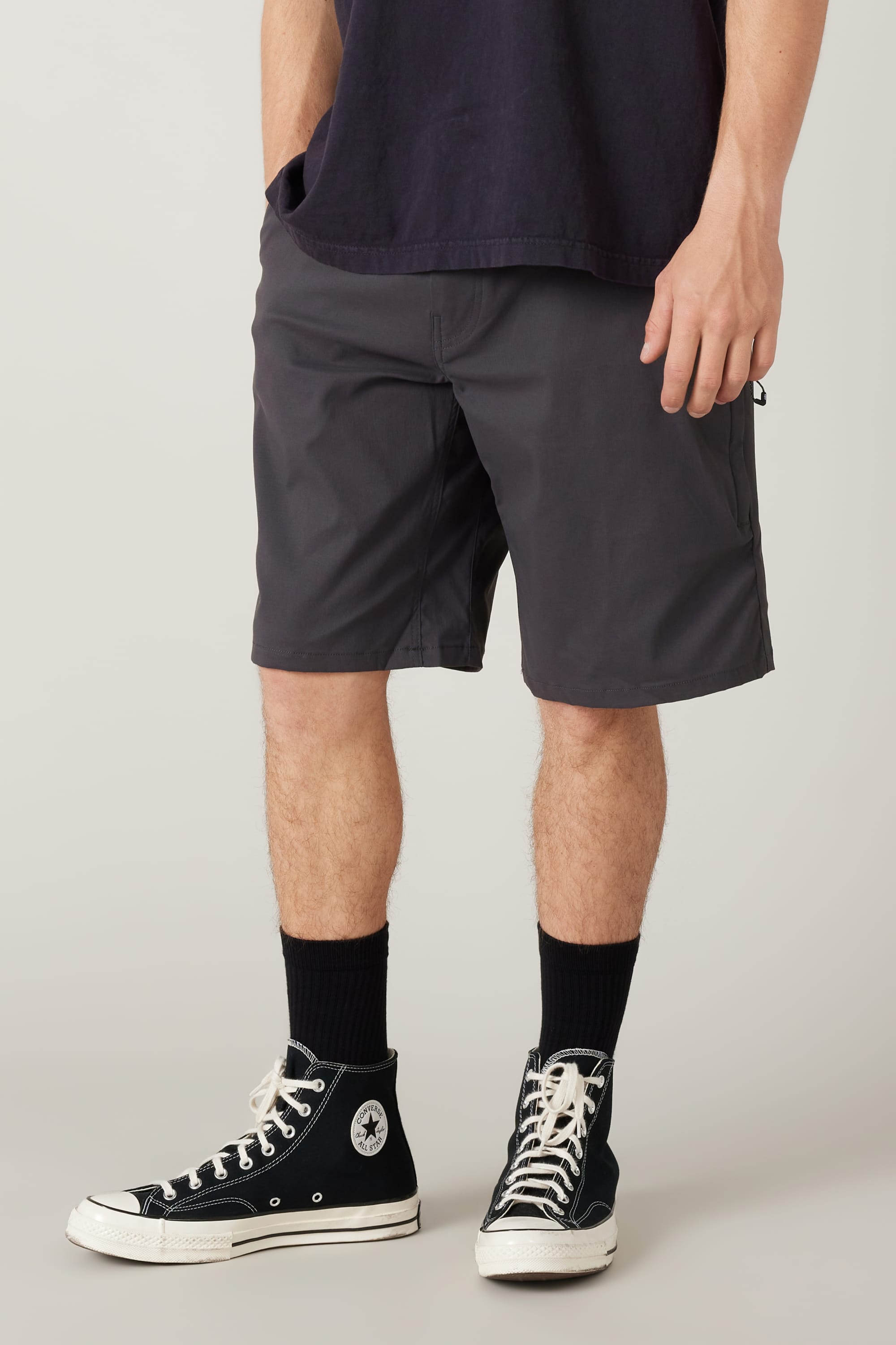 A person wearing a black T-shirt, 686 Men's Everywhere® Hybrid Short - Relaxed Fit in dark gray, black socks, and black Converse high-tops stands against a light background. The image focuses on the person's lower half.