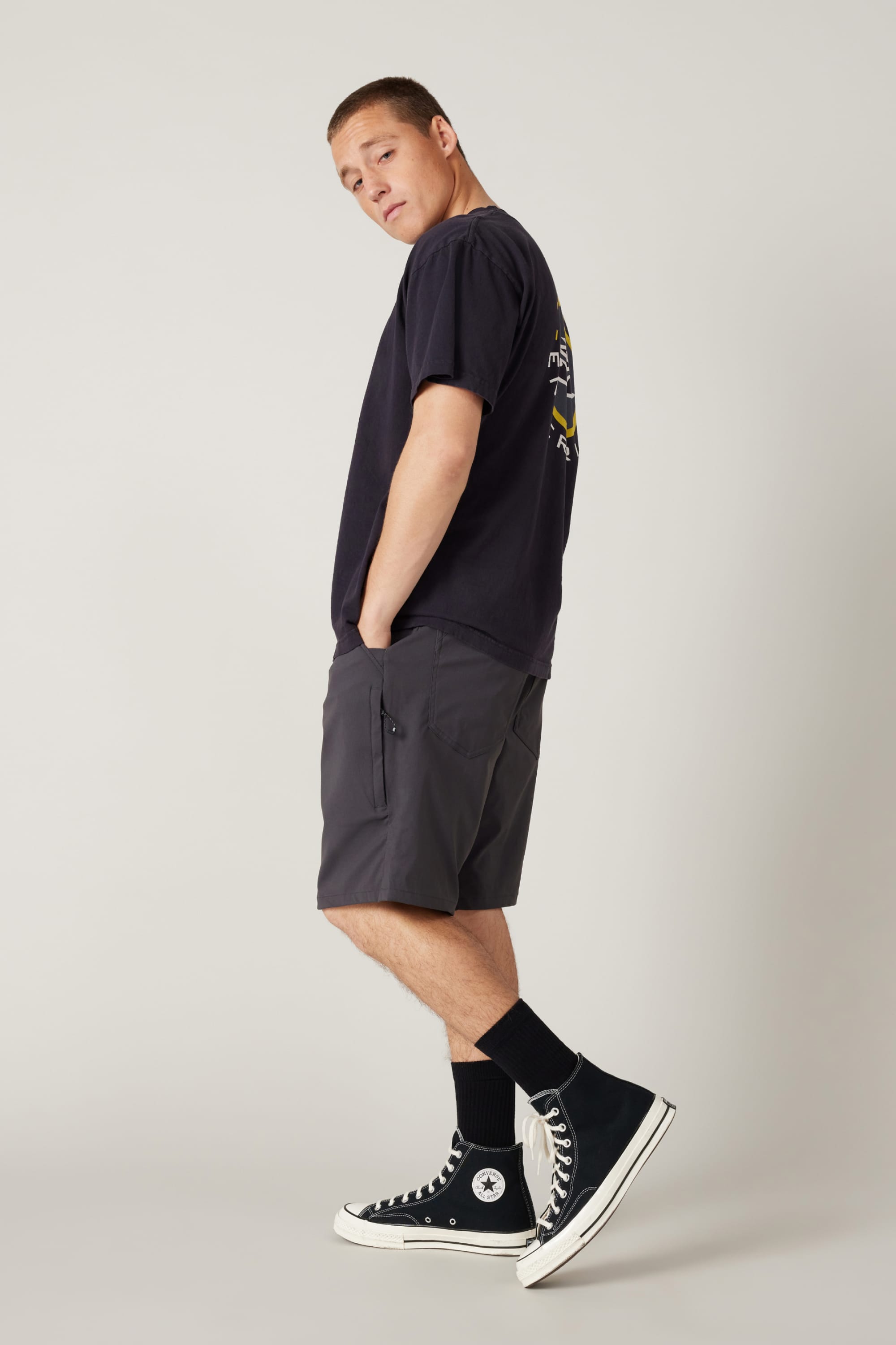 A person stands in profile against a plain background, wearing a black T-shirt, 686 Men's Everywhere® Hybrid Short - Relaxed Fit, black crew socks, and high-top sneakers. Hands in pockets, they look over their shoulder, displaying versatile style.
